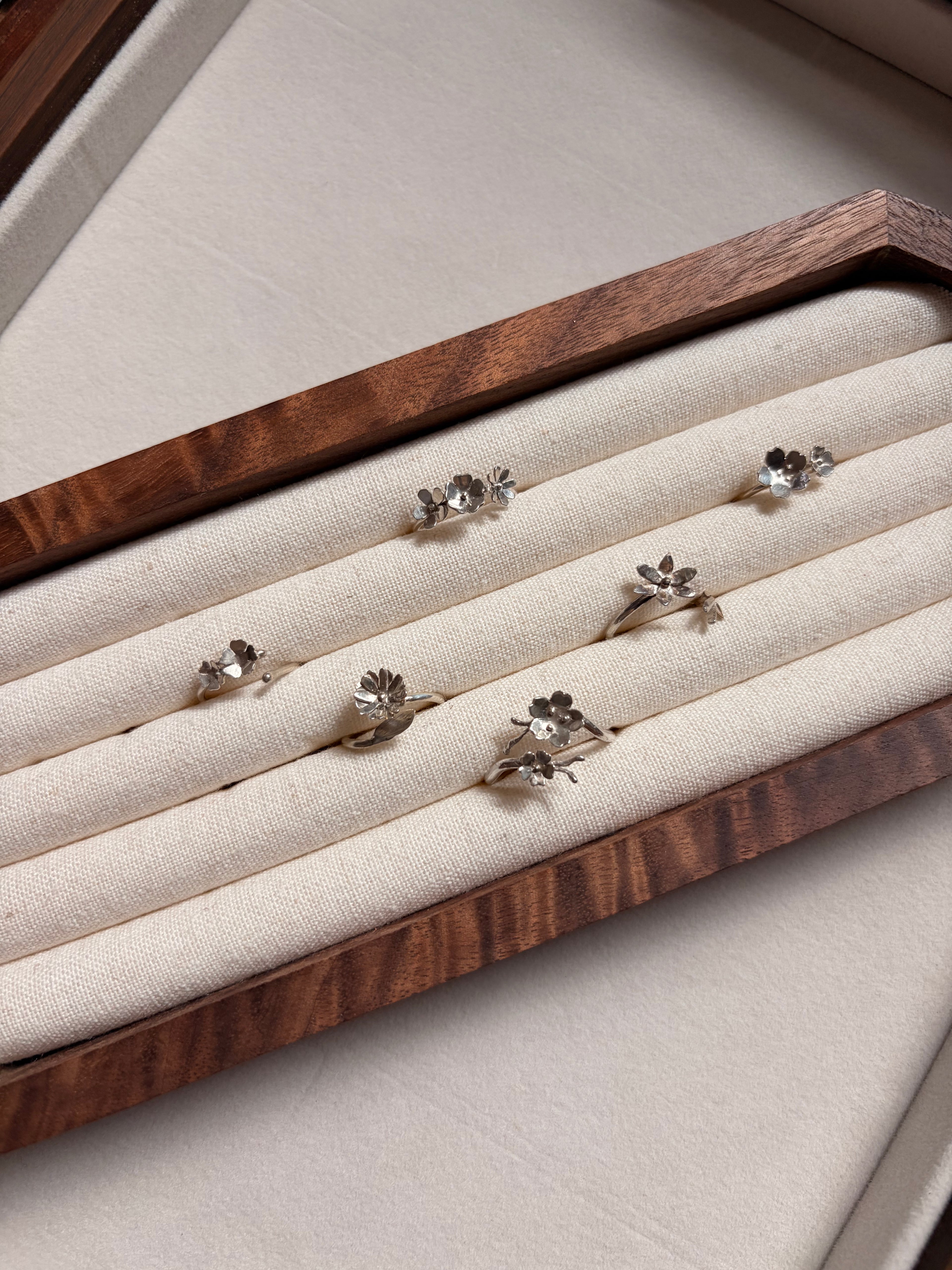 Six silver rings with small flower motifs made in sterling silver and displayed on a cream fabric-lined wooden jewelry box.