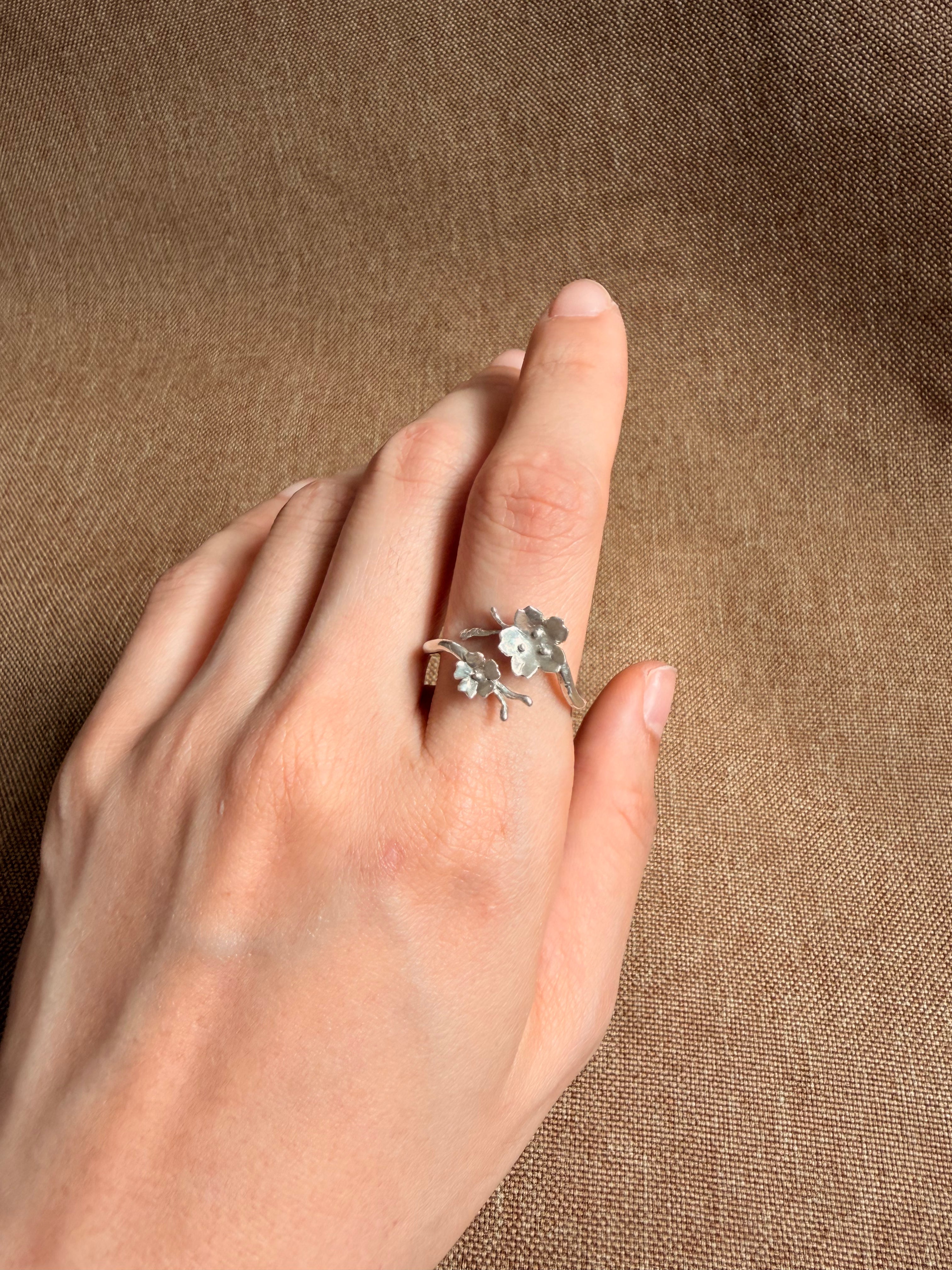 A silver ring with two cherry blossom motifs wrapped around a band, shown worn on a person's finger against a textured brown fabric background.
