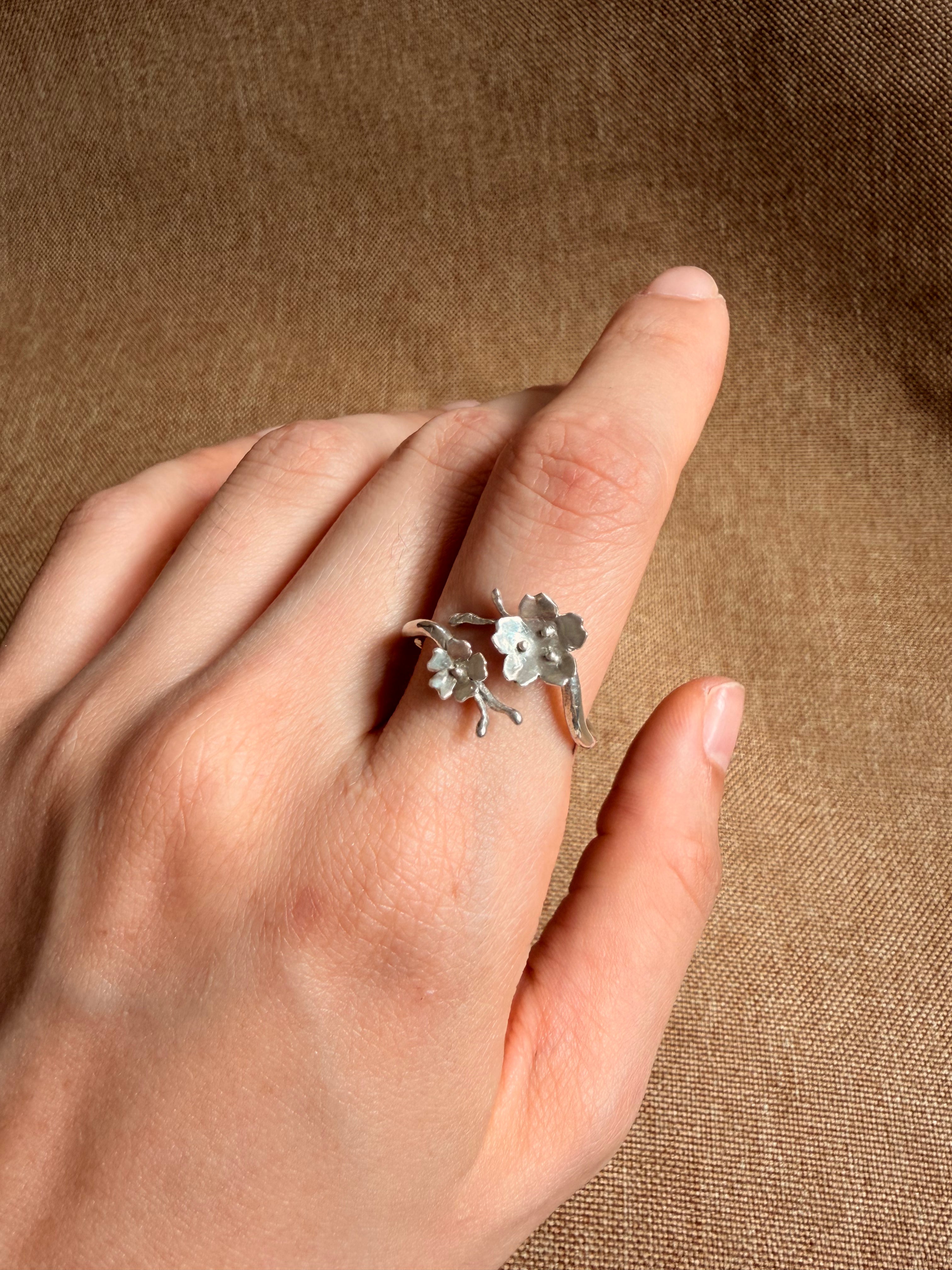 A silver ring with two cherry blossomes charms on a branch, worn on a person's ring finger against a textured brown fabric background.