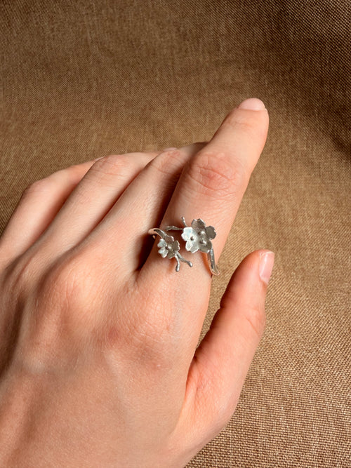 A silver ring with two cherry blossomes charms on a branch, worn on a person's ring finger against a textured brown fabric background.