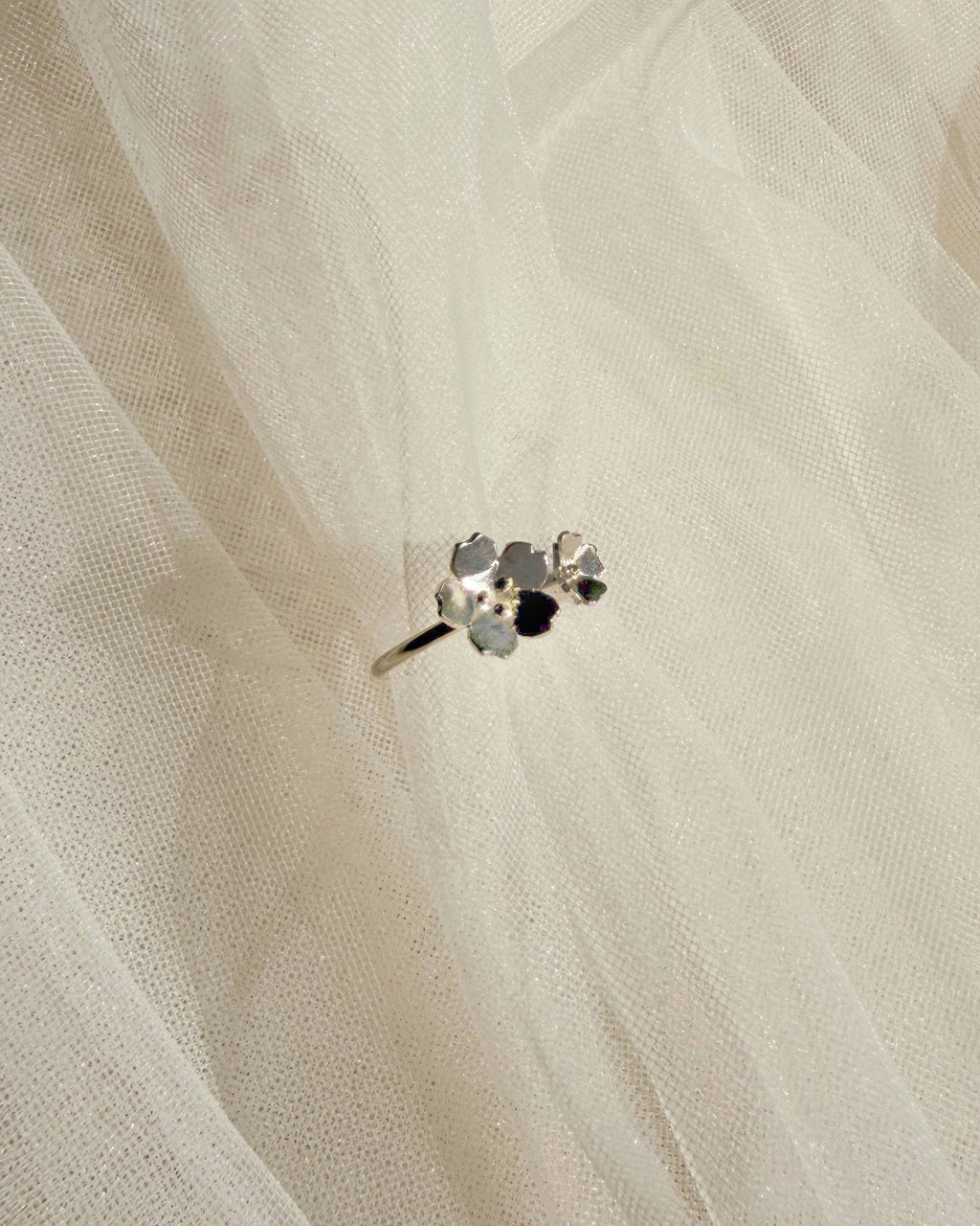 A close-up of a delicate silver-toned ring with small dainty florals set against a soft, sheer white fabric background.