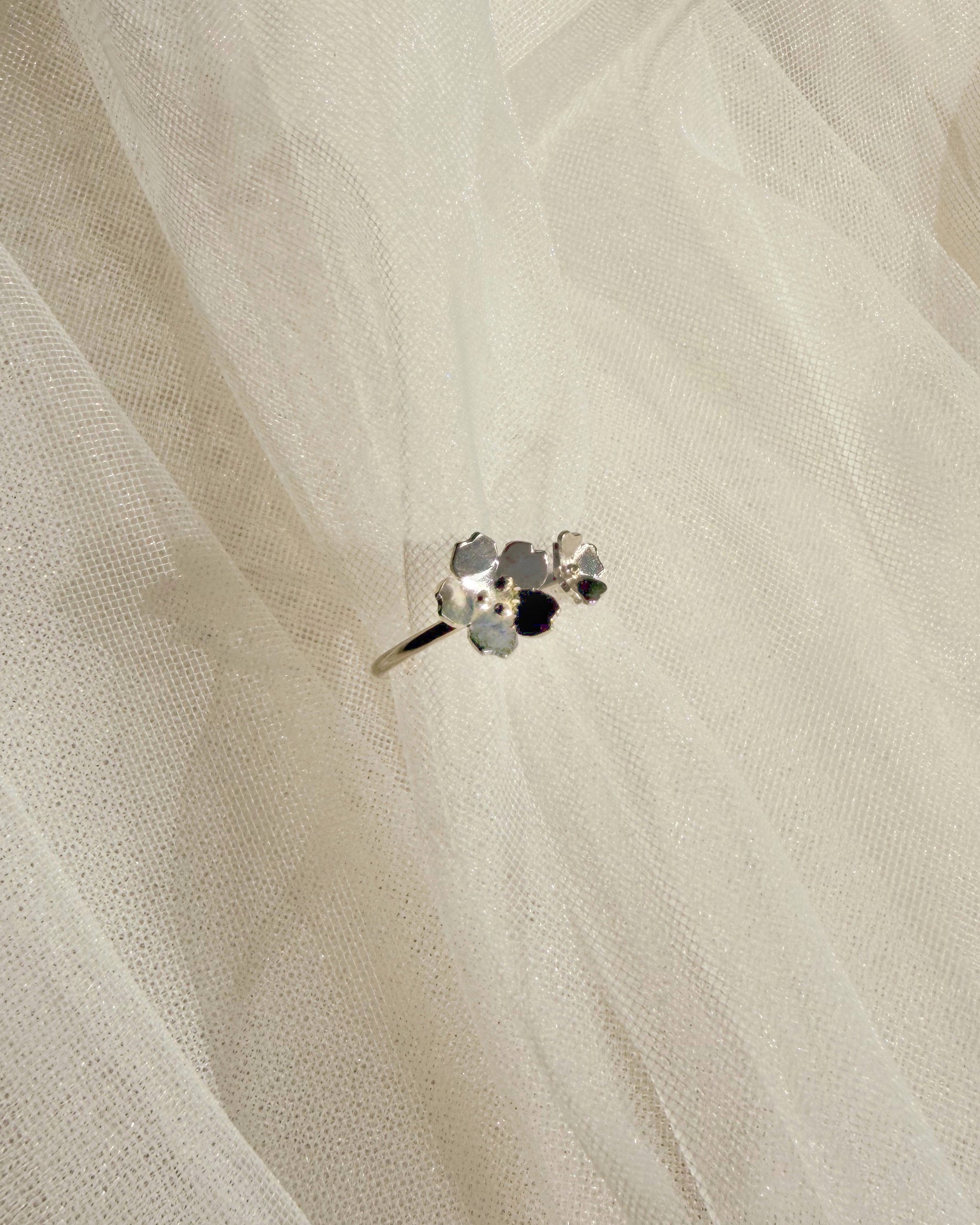 A close-up of a delicate silver-toned ring with small dainty florals set against a soft, sheer white fabric background.