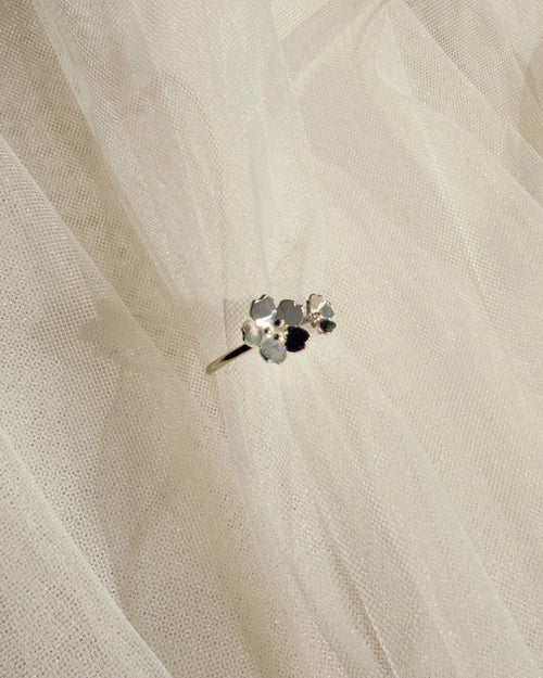 A close-up of a delicate silver-toned ring with small dainty florals set against a soft, sheer white fabric background.