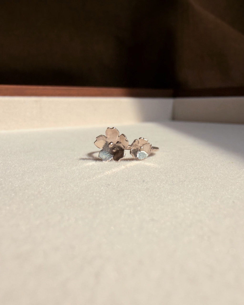 A small silver-toned ring with two floral motifs, resting on a textured off-white surface with a dark brown background.