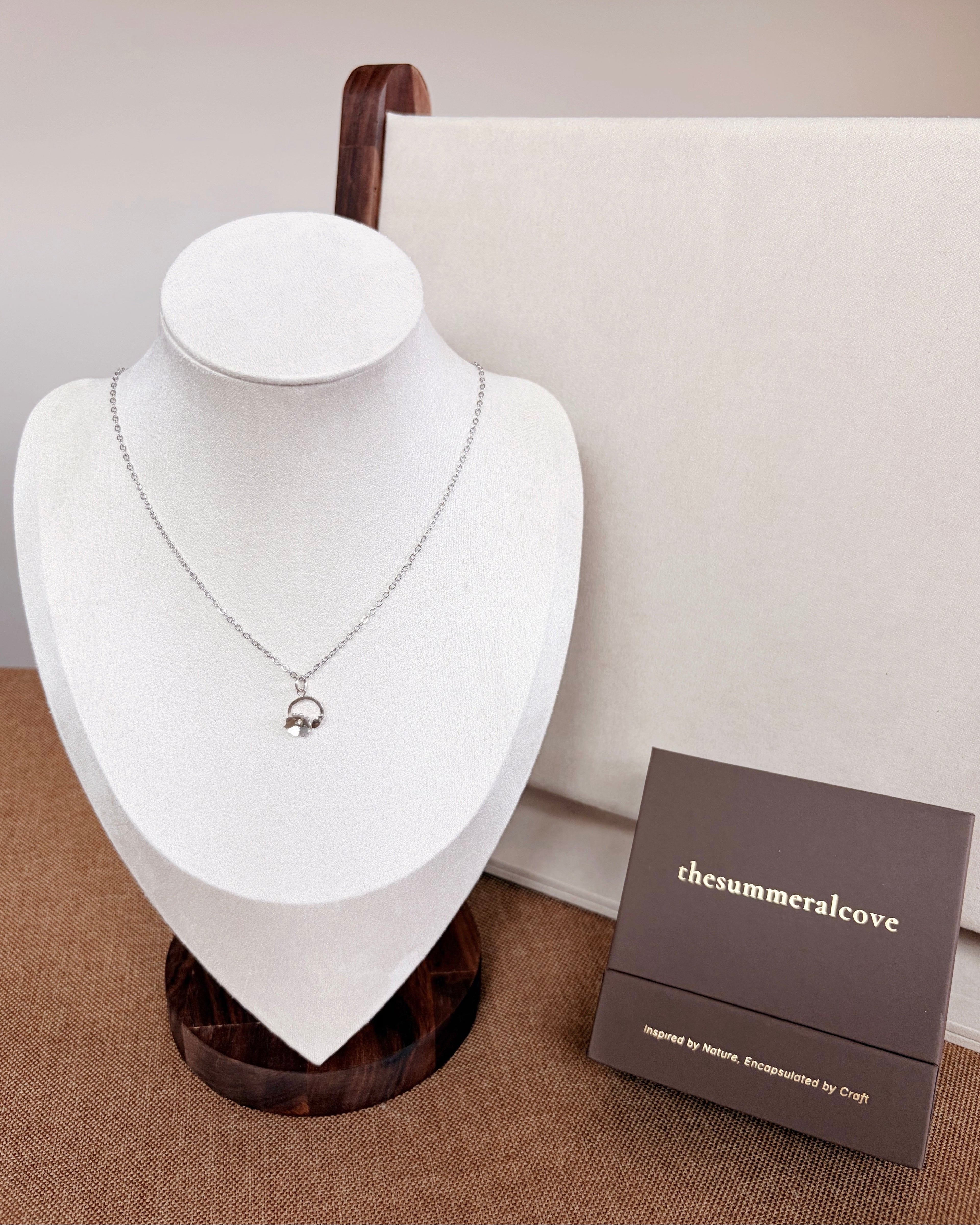 A silver-toned delicate chain necklace with a small hanging floral pendant displayed on a white jewelry bust. A dark brown branded box labeled 'thesummeralcove' sits beside the bust on a textured brown surface against a neutral beige background.