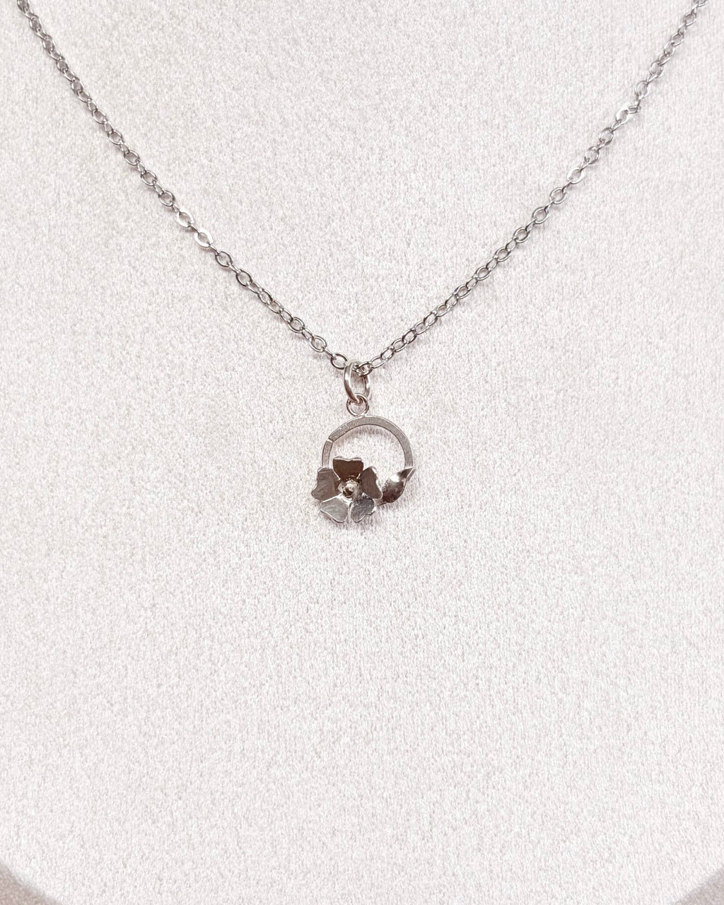 A delicate silver chain necklace with a small circular floral pendant displayed on a white jewelry bust against a neutral indoor background.