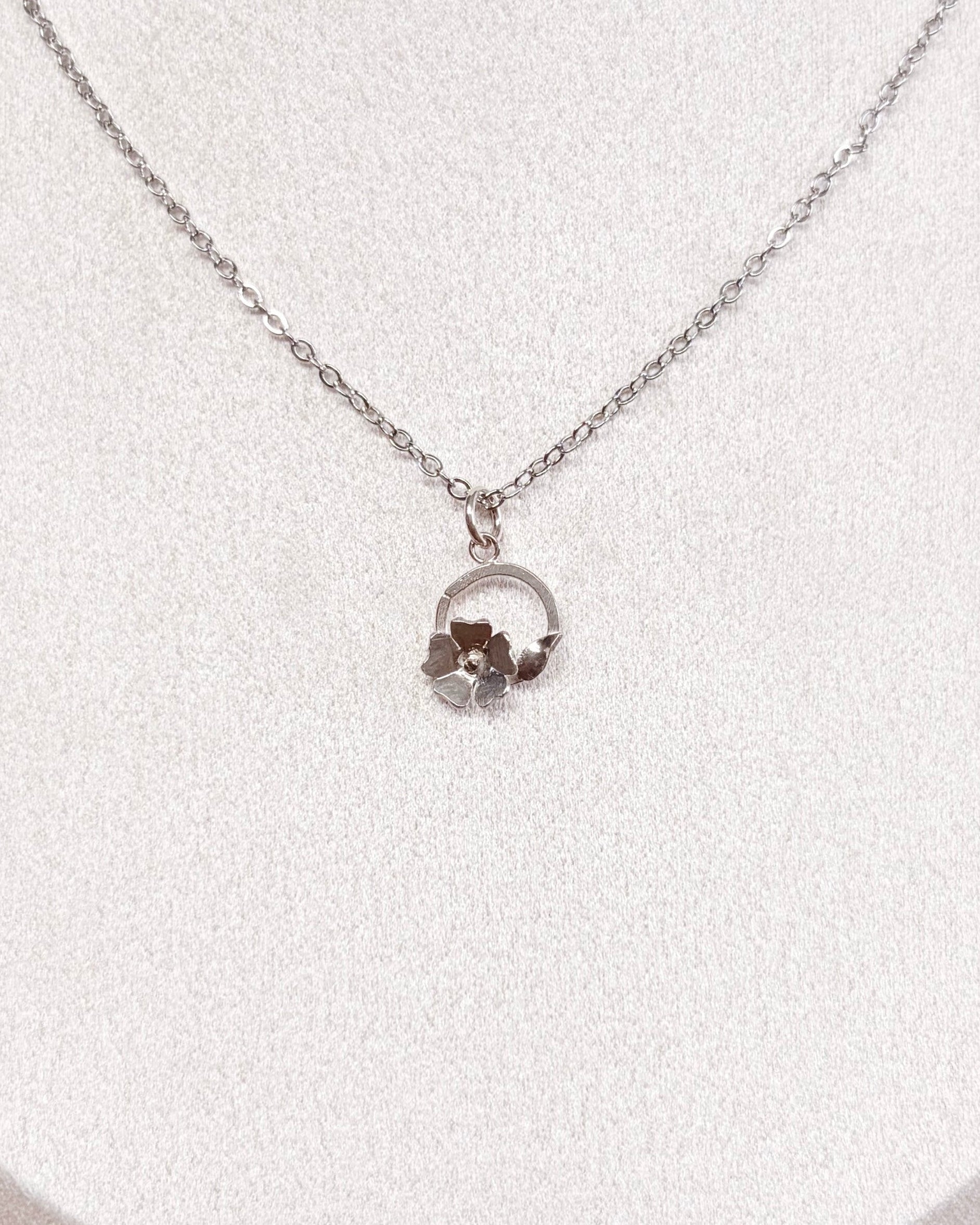 A delicate silver chain necklace with a small circular floral pendant displayed on a white jewelry bust against a neutral indoor background.