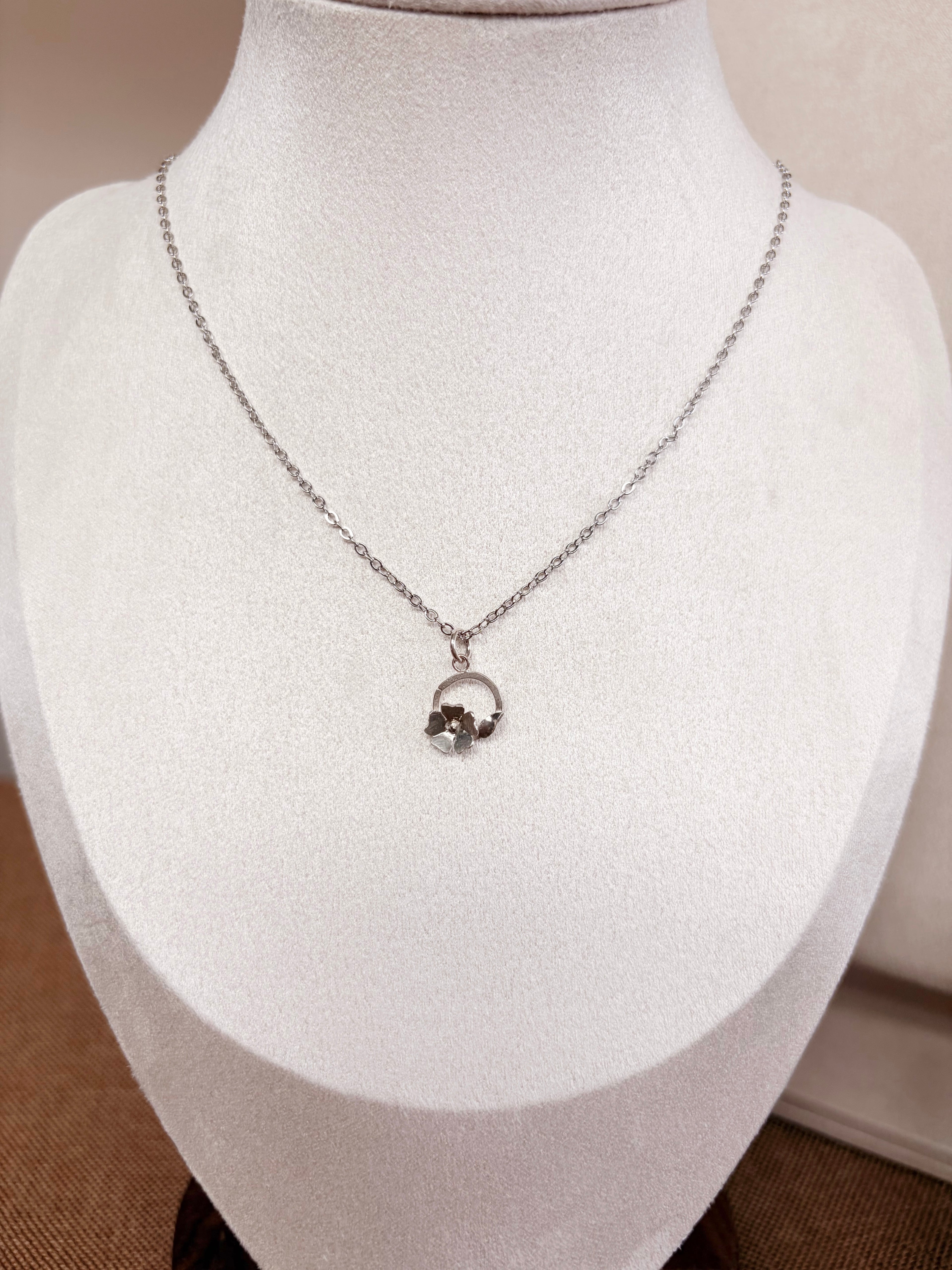 A silver chain necklace with a small circular floral pendant , displayed on a white jewelry bust.