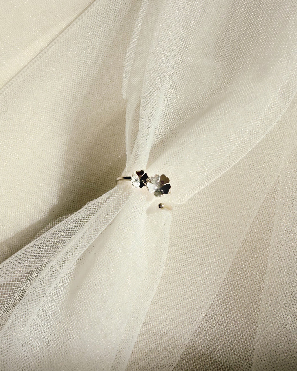 A close-up of a white tulle or mesh fabric with a  silver floral ring near the center, set against draped folds of the same translucent white material.
