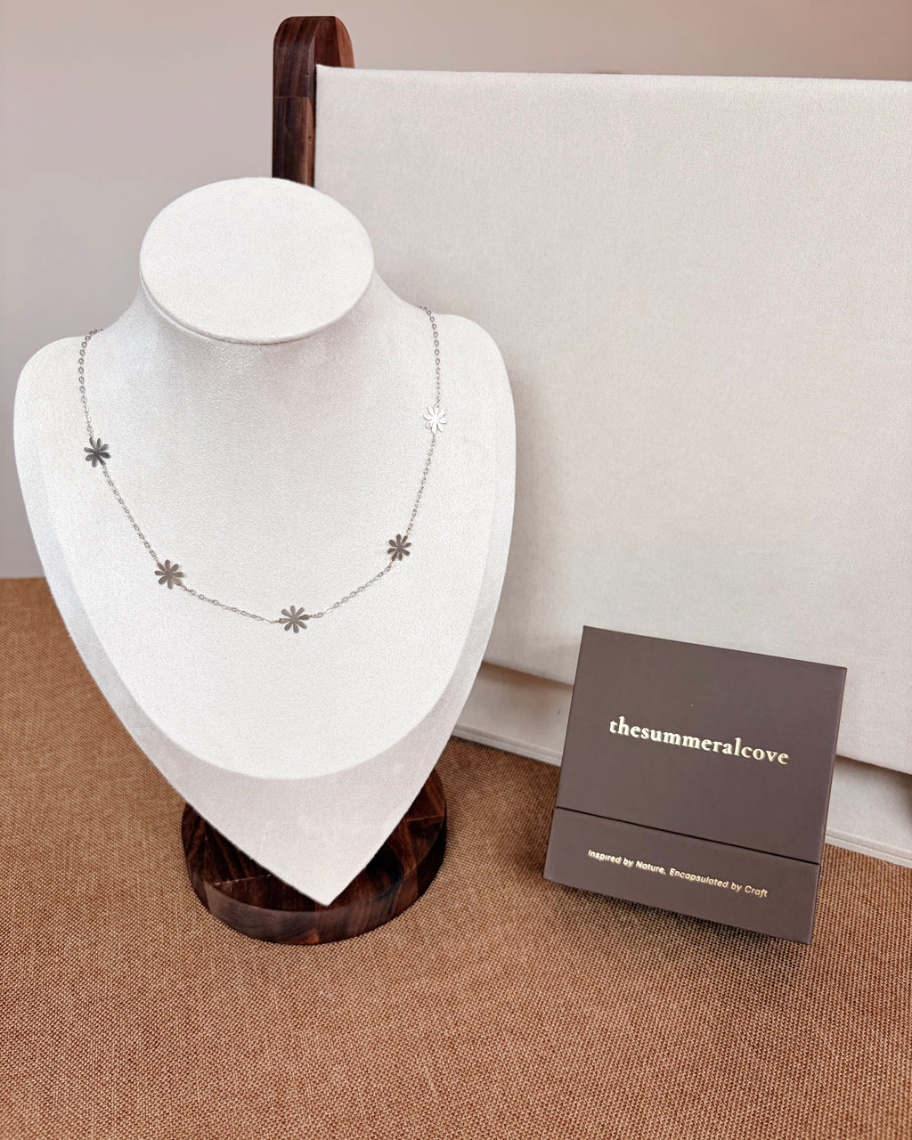 A delicate silver necklace with small daisy charms displayed on a white jewelry bust, with a dark brown brand box labeled 'thesummeralcove' beside it on a textured beige surface.