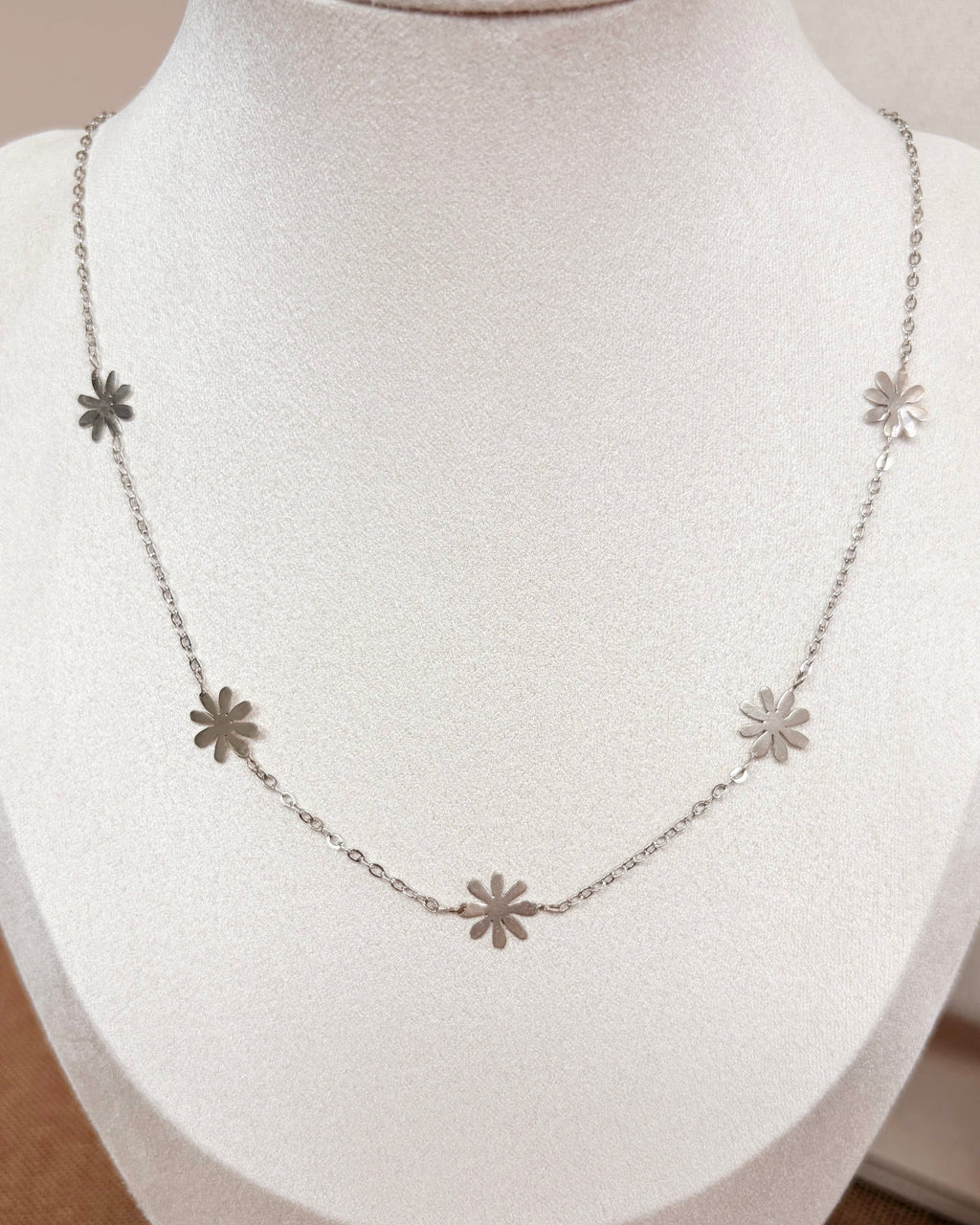 A delicate silver necklace with five small daisy flower-shaped charms spaced along the chain, displayed on a white fabric jewelry bust.