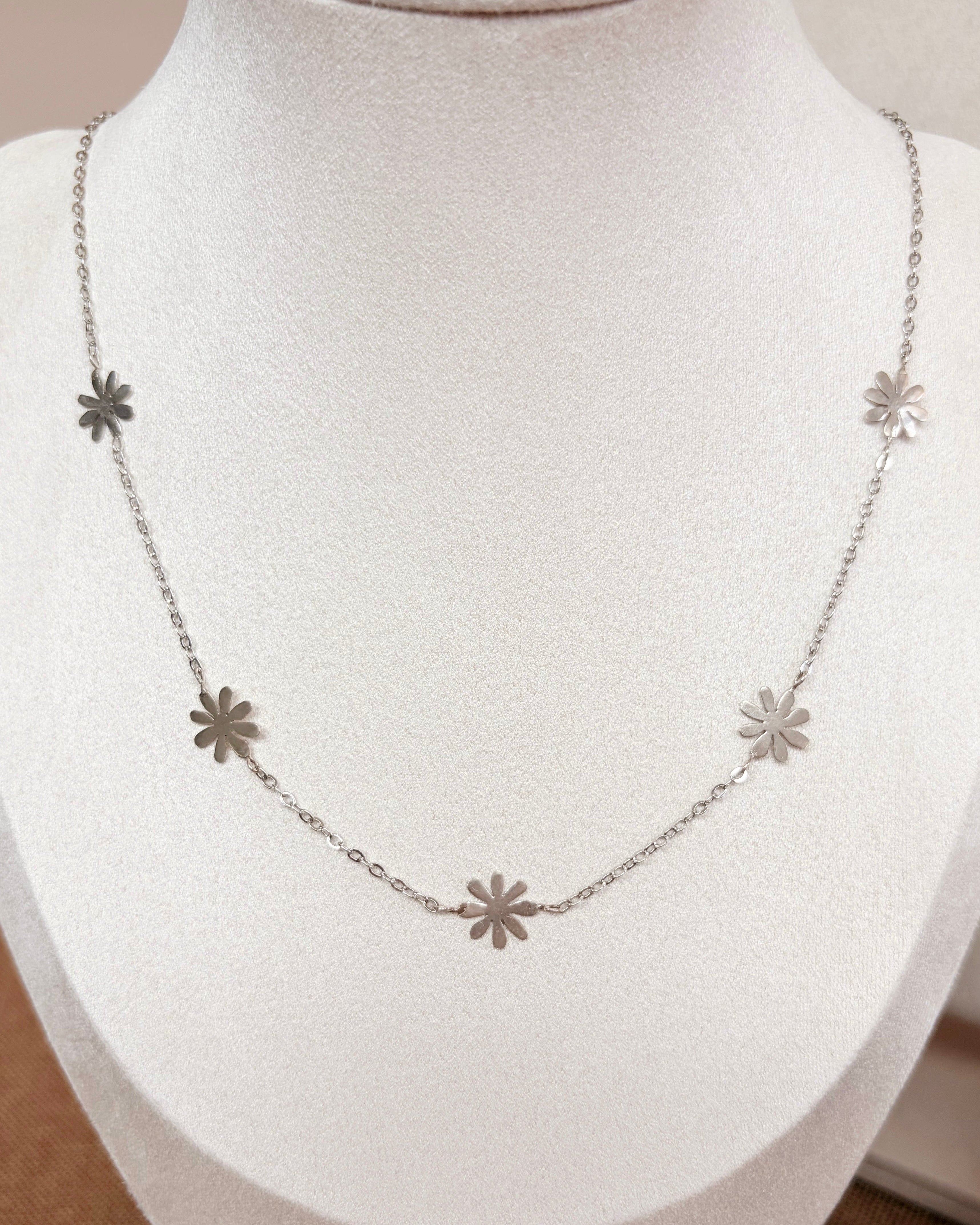 A delicate silver necklace with five small daisy flower-shaped charms spaced along the chain, displayed on a white fabric jewelry bust.
