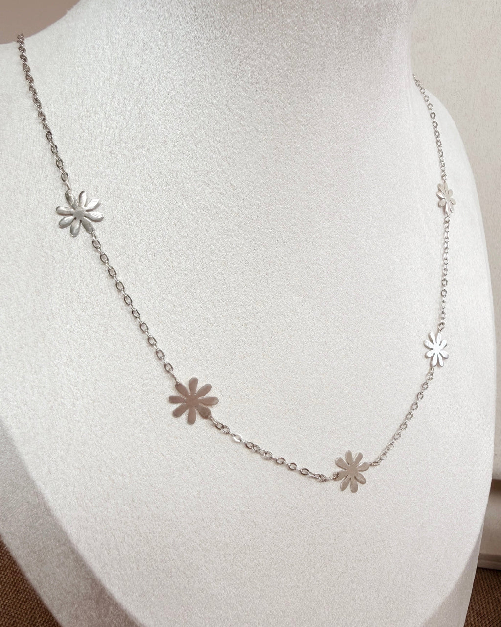 A delicate silver chain necklace with five small flower-shaped charms, displayed on a white jewelry bust.