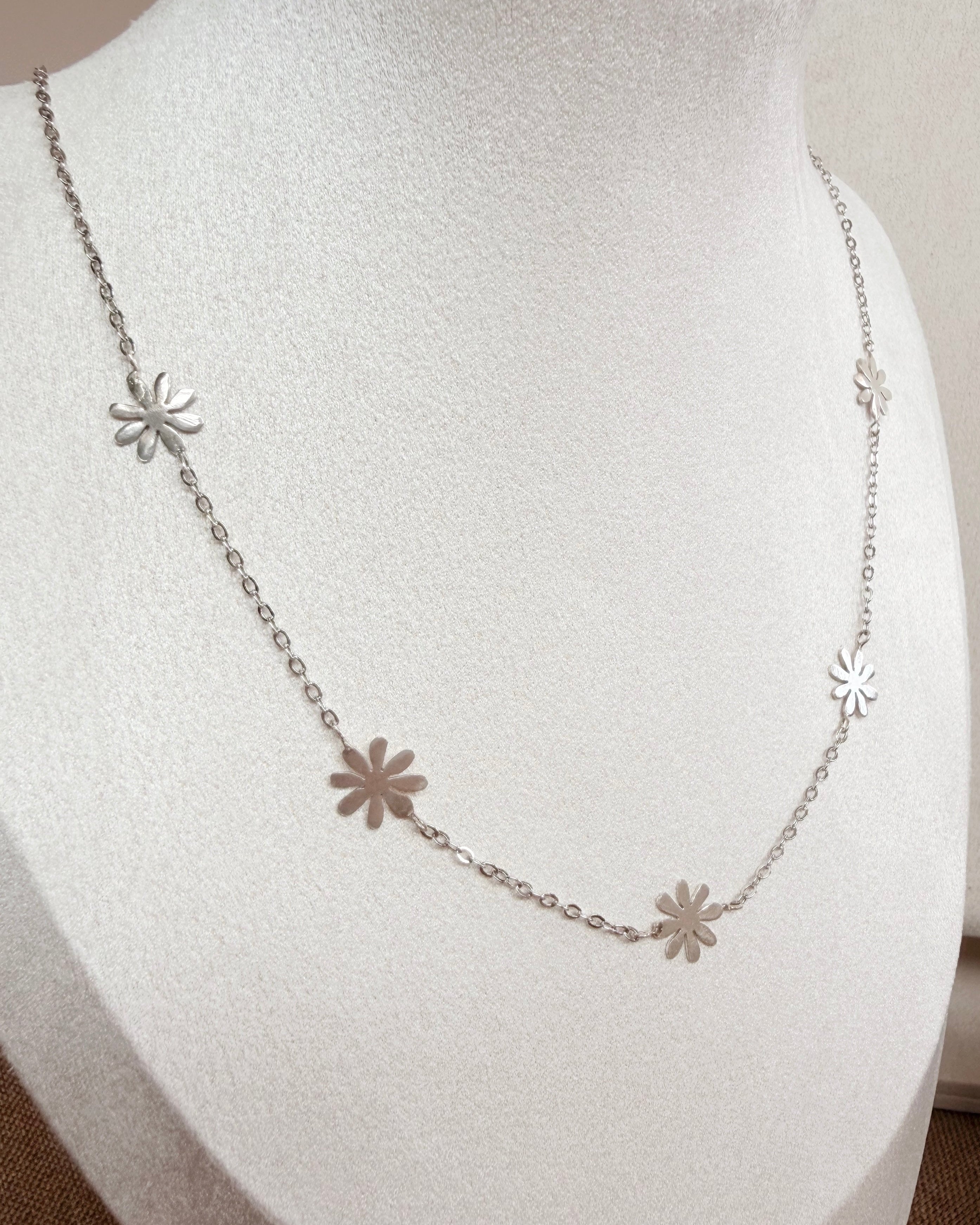A delicate silver chain necklace with five small flower-shaped charms, displayed on a white jewelry bust.