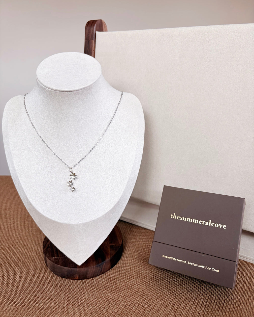 A delicate silver necklace with a small floral pendant displayed on a white jewelry bust. A dark brown brand box labeled 'thesummeralcove' sits beside the bust on a textured tan surface, with a cream backdrop behind.