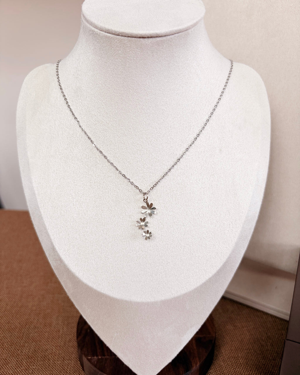 A delicate silver necklace with a small floral pendant displayed on a white fabric jewelry bust against a neutral indoor background.