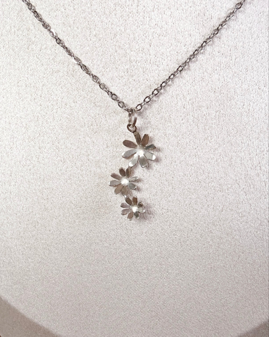 A silver daisy necklace with a delicate chain and three small flower charms, displayed on a white textured jewelry bust.