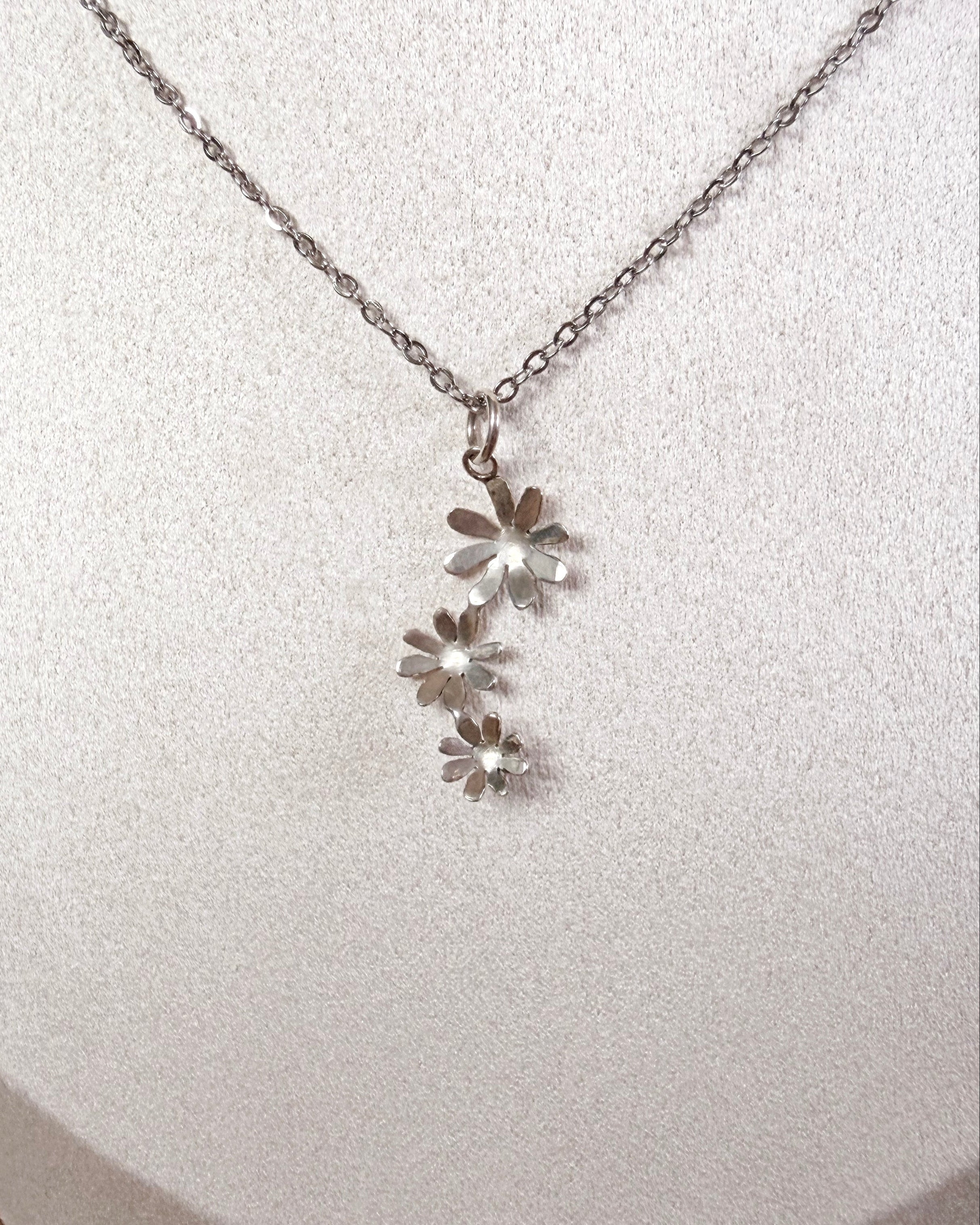 A silver daisy necklace with a delicate chain and three small flower charms, displayed on a white textured jewelry bust.