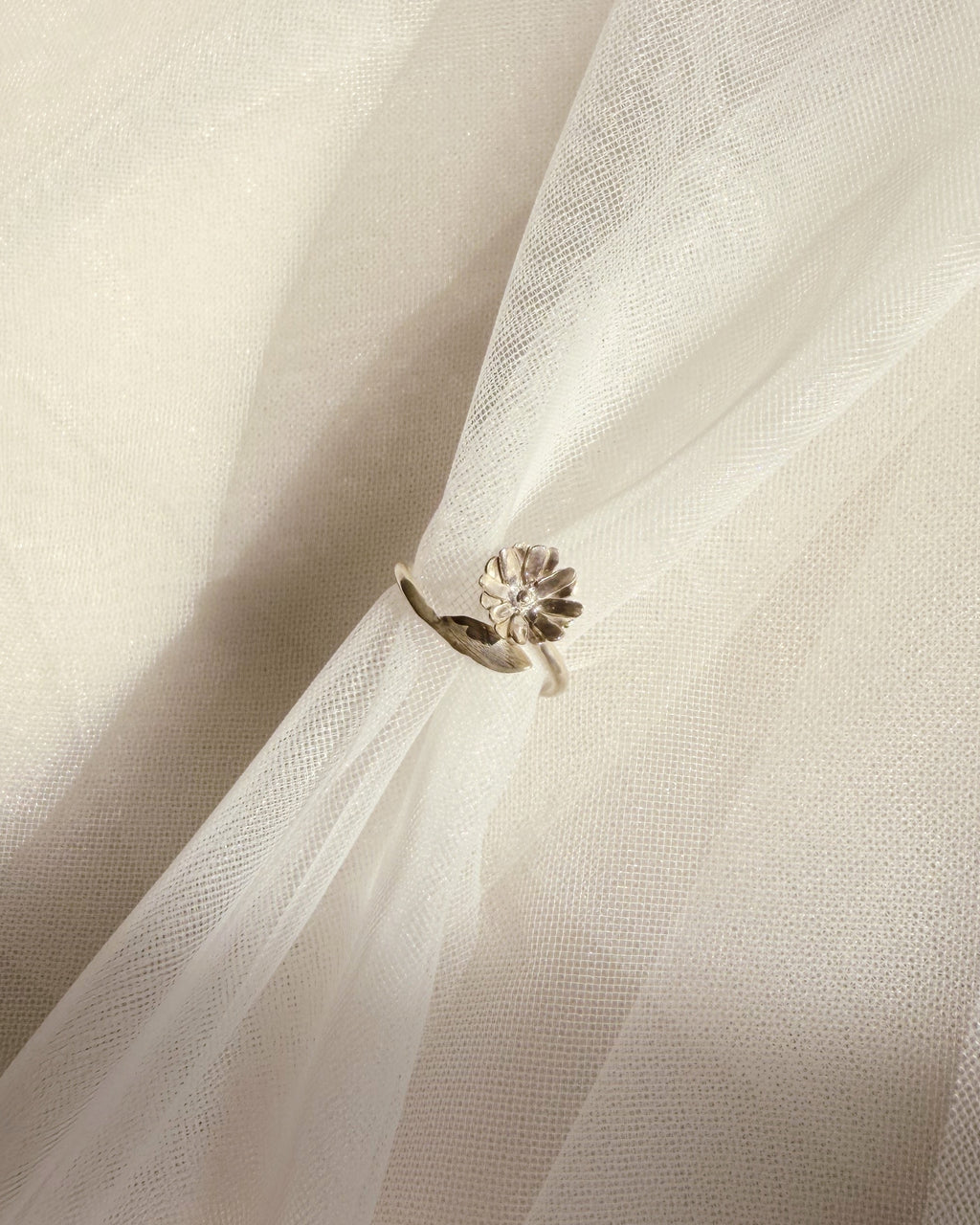 A silver-toned ring with a small floral and leaf-like cluster setting, shown resting on draped white tulle fabric.