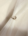 A silver-toned ring with a small floral and leaf-like cluster setting, shown resting on draped white tulle fabric.