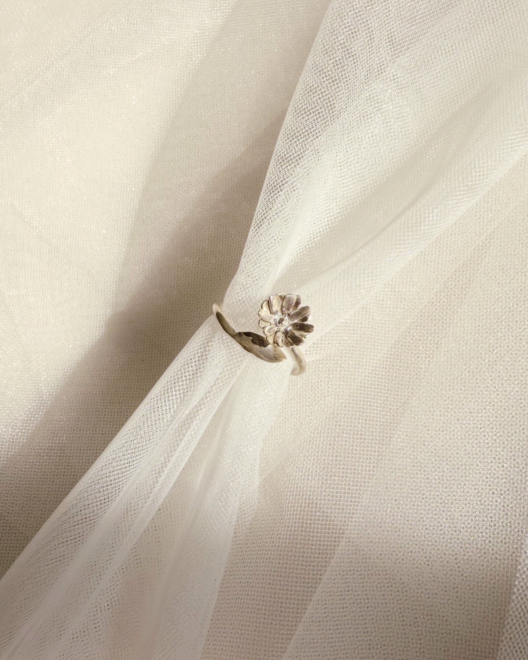 A silver-toned ring with a small floral and leaf-like cluster setting, shown resting on draped white tulle fabric.