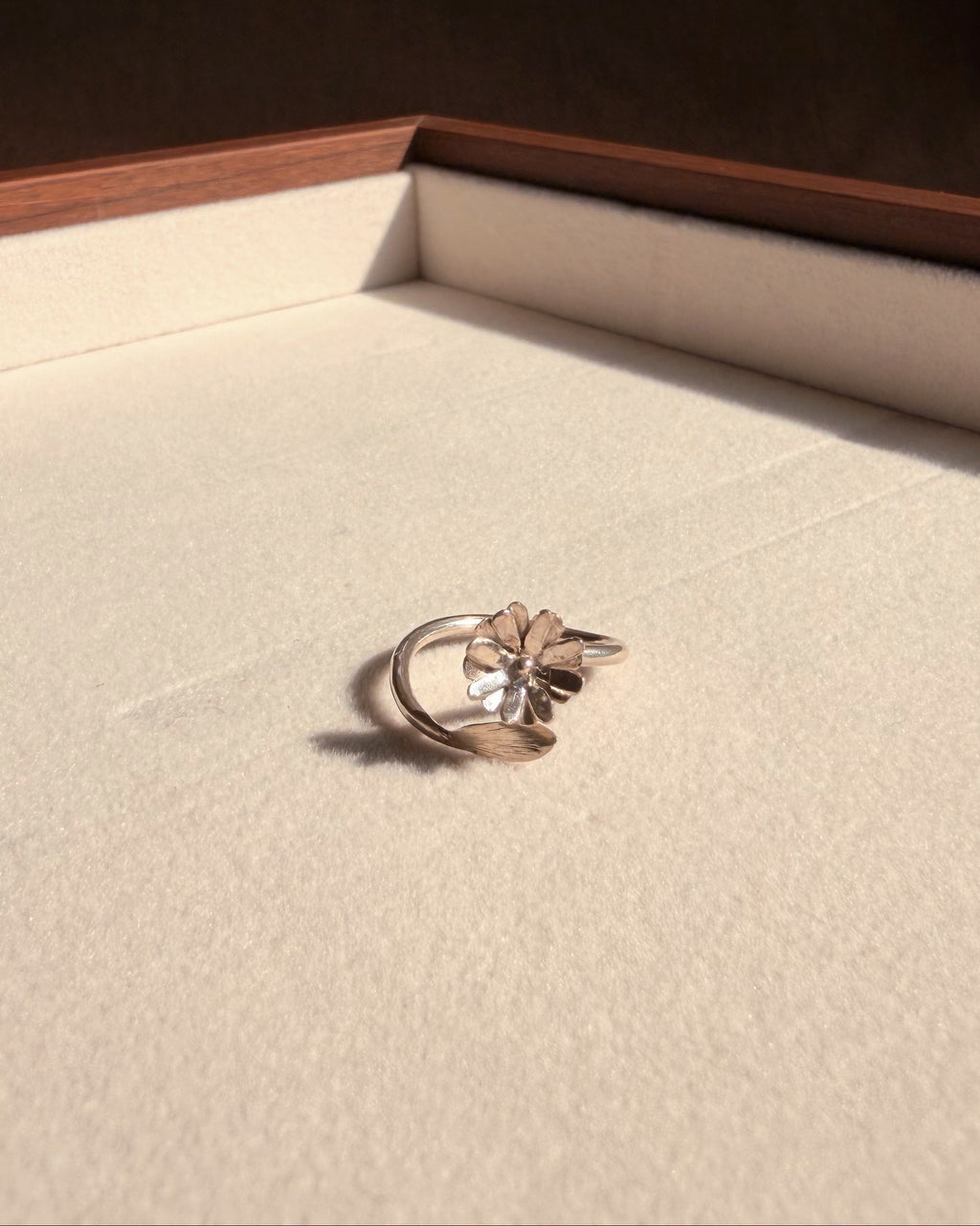 A silver-toned ring with a clear central flower and curved side details, resting on a cream-colored jewelry box insert.