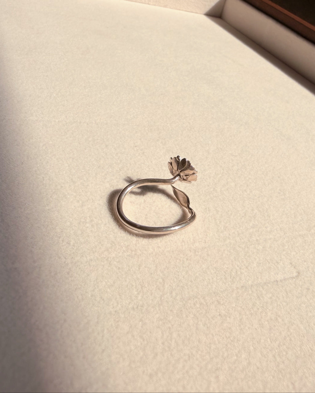 A silver ring with a small flower-shaped accent on the band, resting on a textured cream-colored surface inside a box.