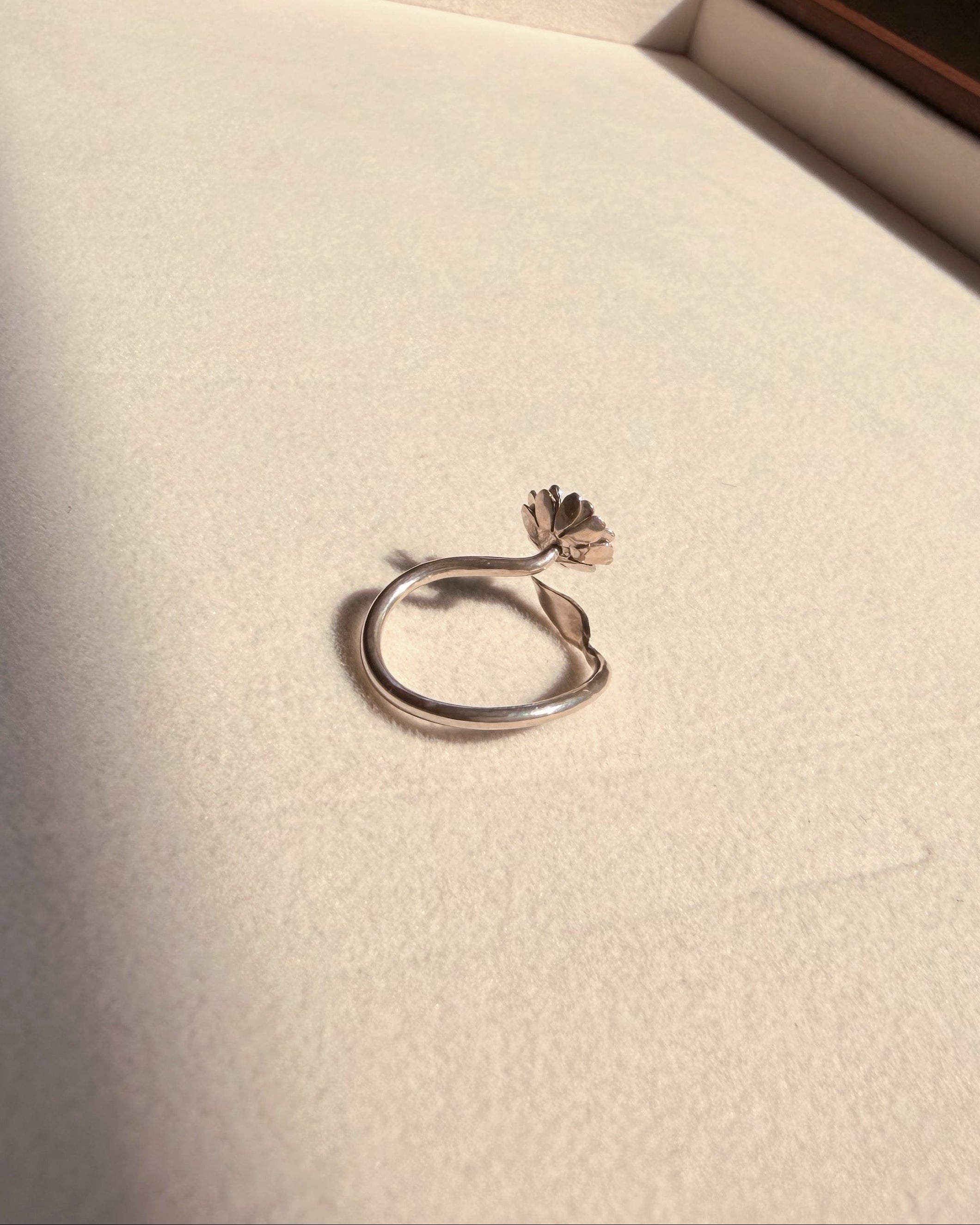 A silver ring with a small flower-shaped accent on the band, resting on a textured cream-colored surface inside a box.