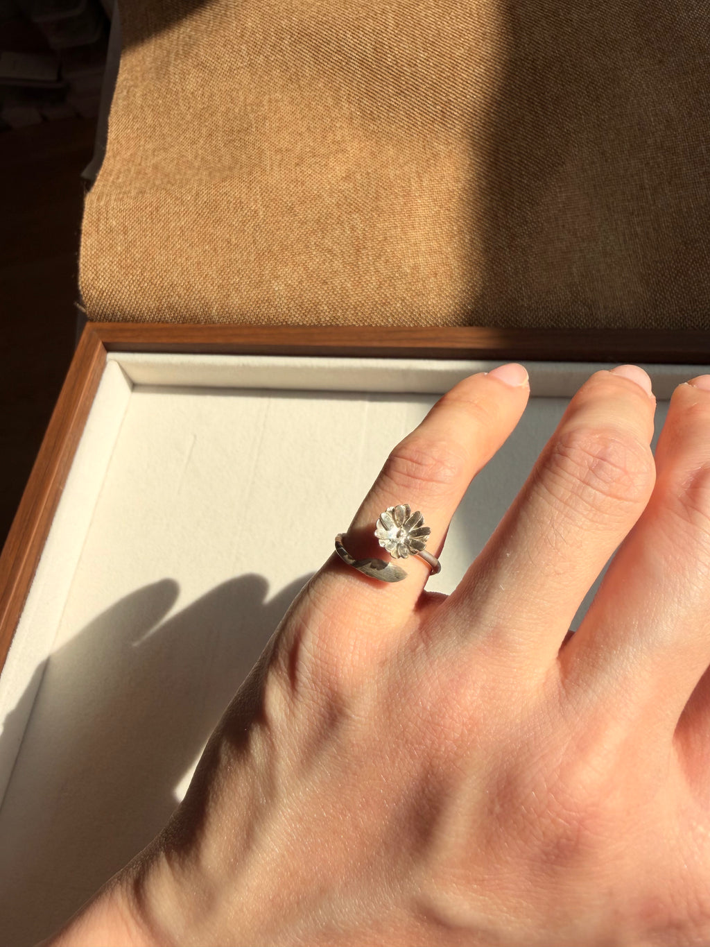 A silver flower ring with a dandelion motif worn on a person's pinky finger, shown against a warm brown and beige background.