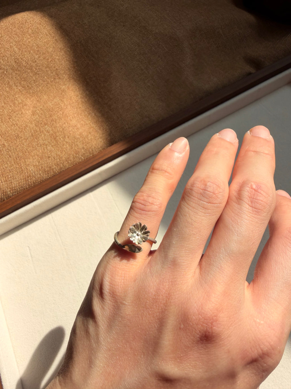 A close-up of a hand wearing a silver ring with a dandelion flower and leaf, set against a neutral background with a textured brown box.