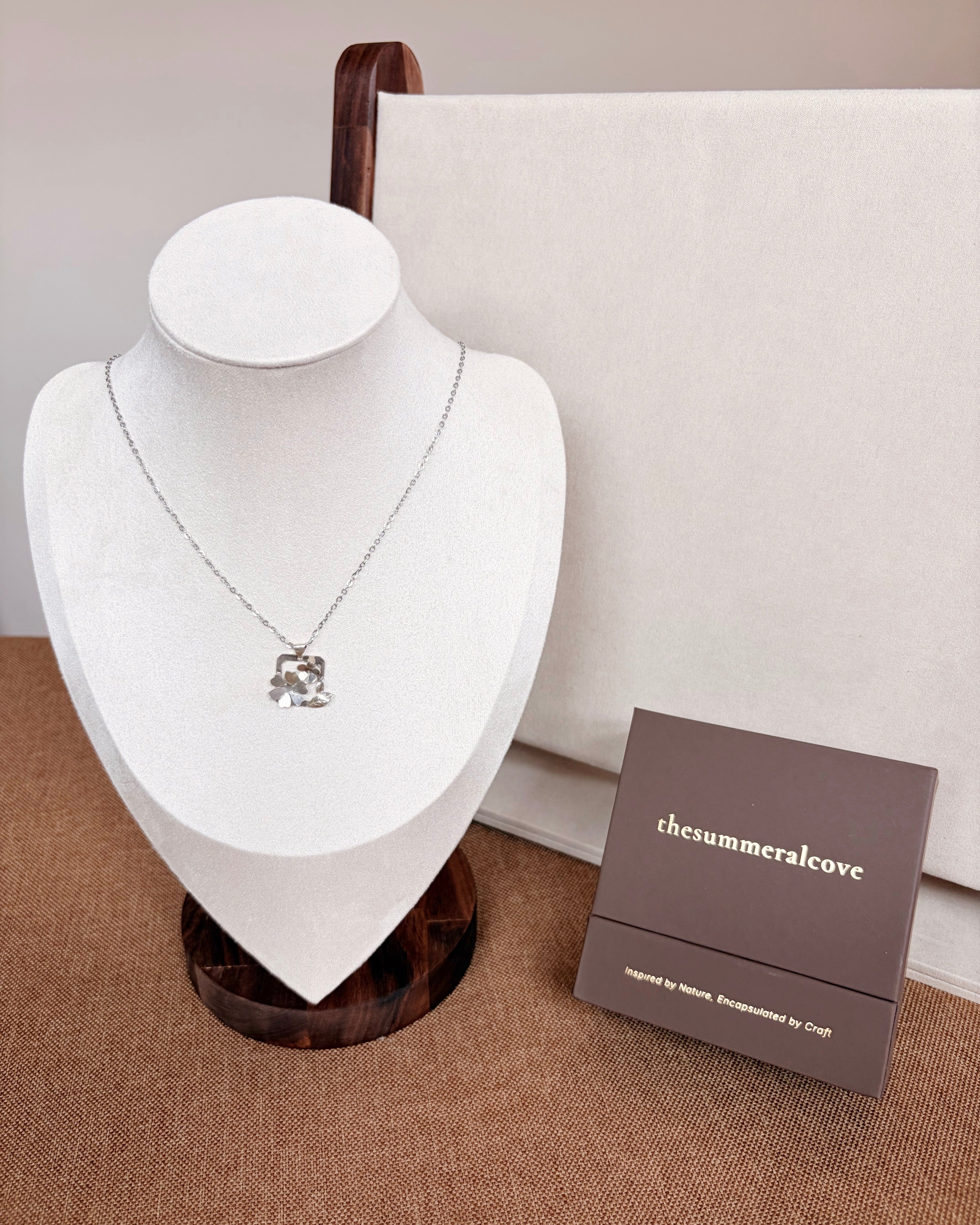 A silver-toned necklace with a small floral pendant displayed on a white jewelry bust, with a brown presentation box labeled 'thesummeralcove' beside it on a beige surface.
