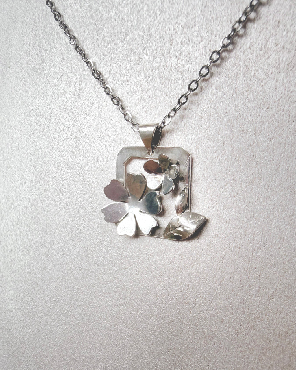 A silver necklace with a geometric floral pendant featuring layered metallic leaves and petals, displayed on a textured light gray bust.