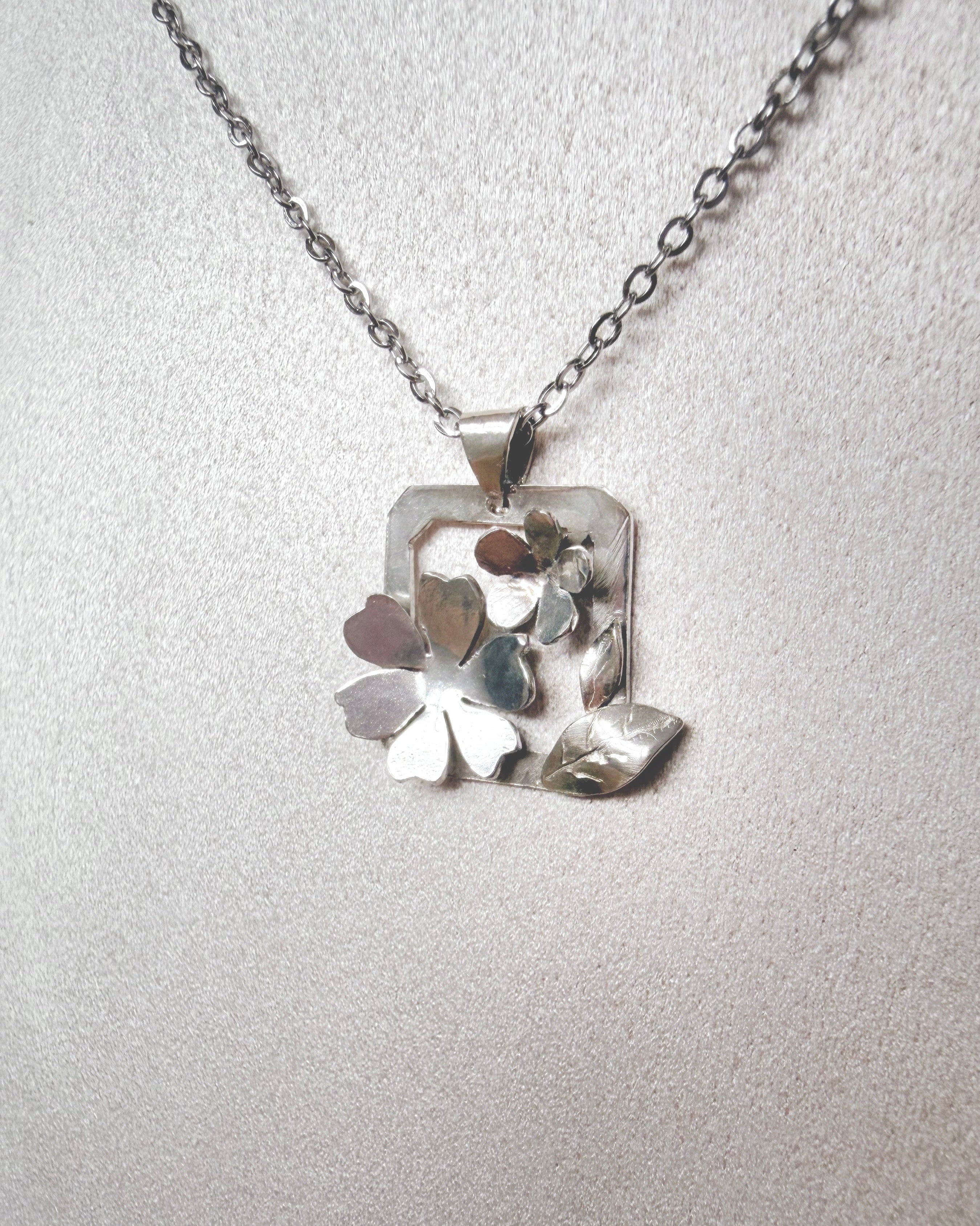 A silver necklace with a geometric floral pendant featuring layered metallic leaves and petals, displayed on a textured light gray bust.