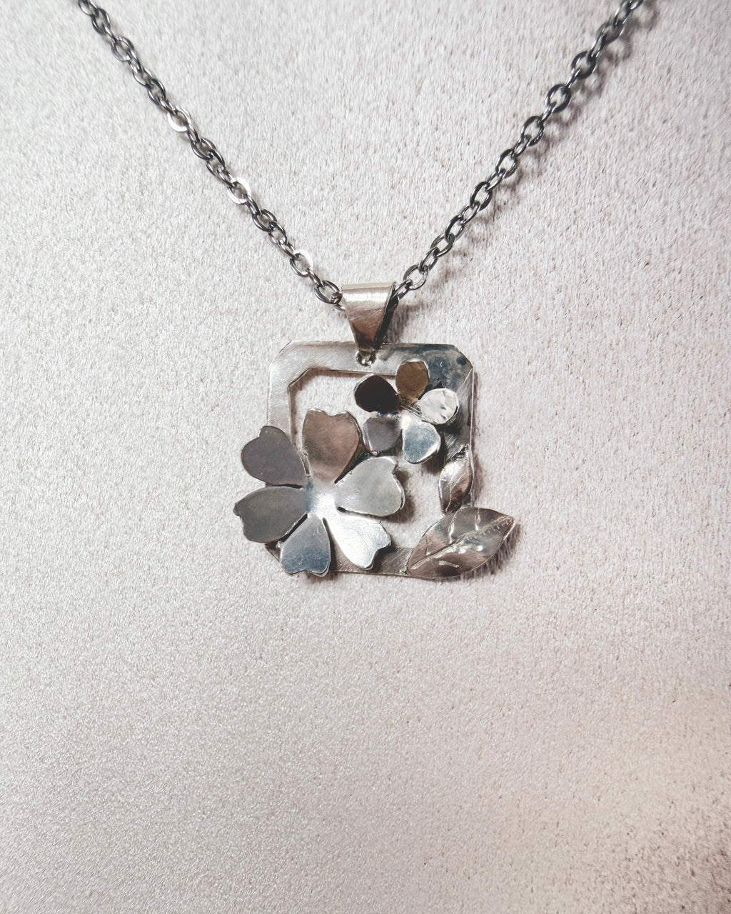 A silver-toned necklace with a square pendant featuring layered flower and leaf shapes, displayed on a textured light beige surface.