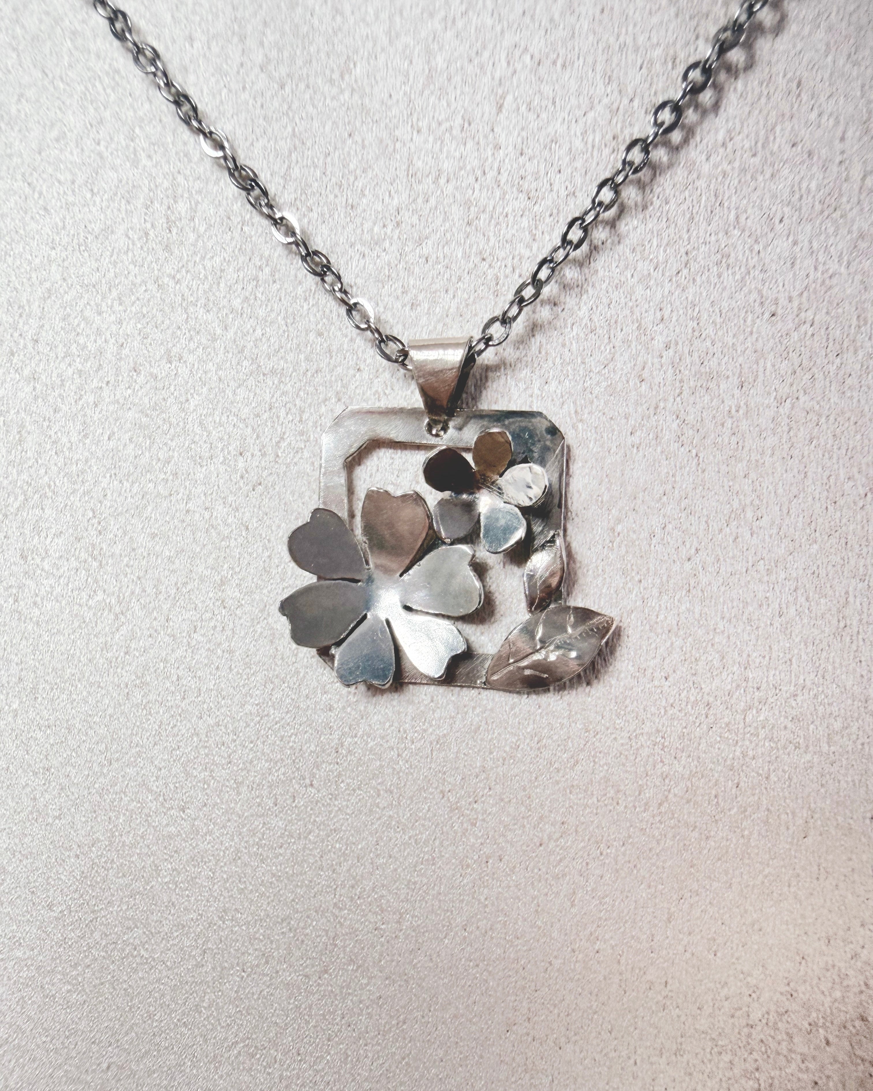 A silver-toned necklace with a square pendant featuring layered flower and leaf shapes, displayed on a textured light beige surface.