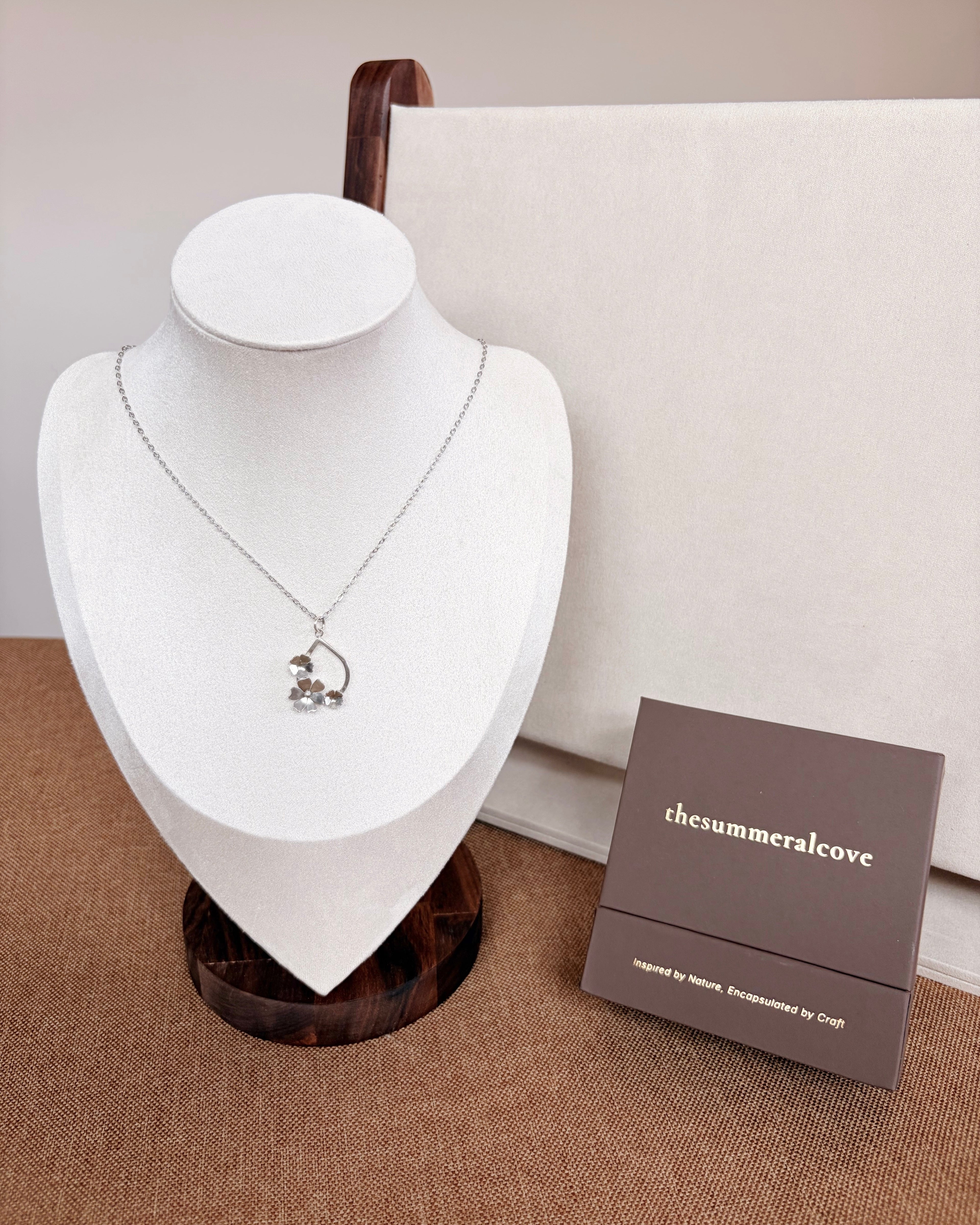 A silver-toned necklace with a teardrop shaped floral pendant displayed on a white jewelry bust. A brown jewelry box labeled 'thesummeralcove' sits beside it on a textured tan surface, with a cream fabric backdrop behind.