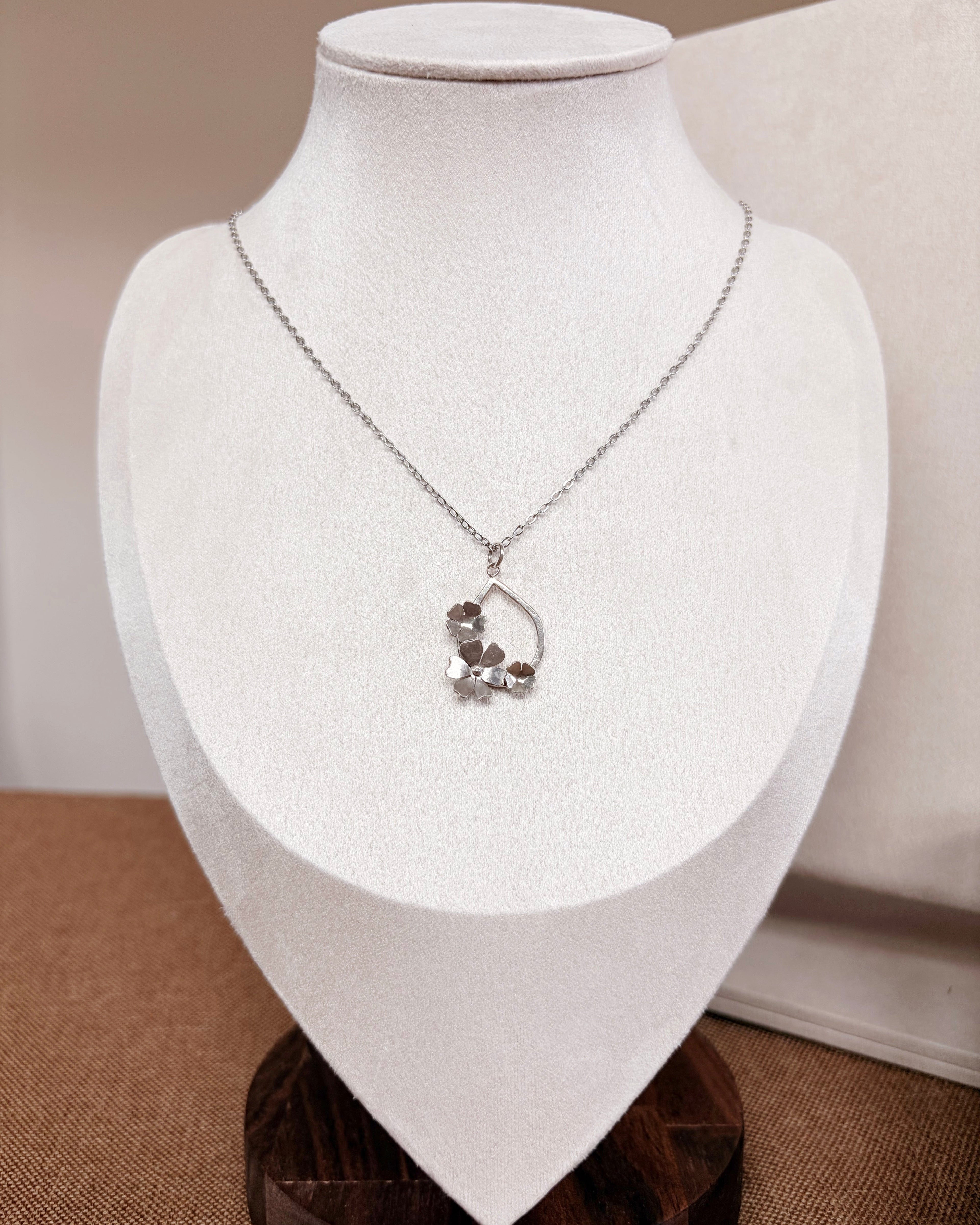 A delicate silver necklace with a small floral pendant displayed on a white fabric jewelry bust.