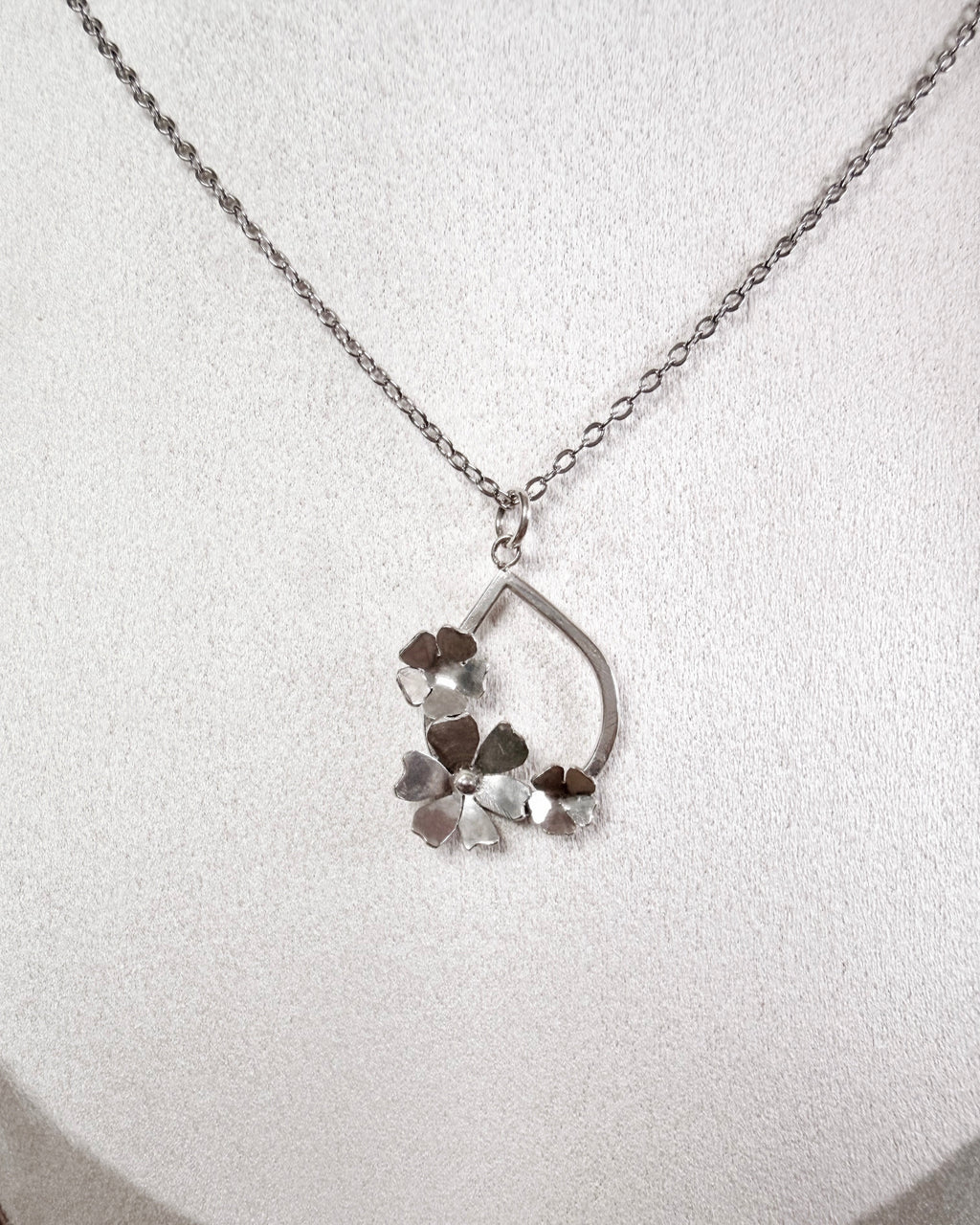 A silver necklace with a delicate chain and a pendant featuring a teardrop-shaped frame and three small flower-like charms, displayed on a textured white surface.