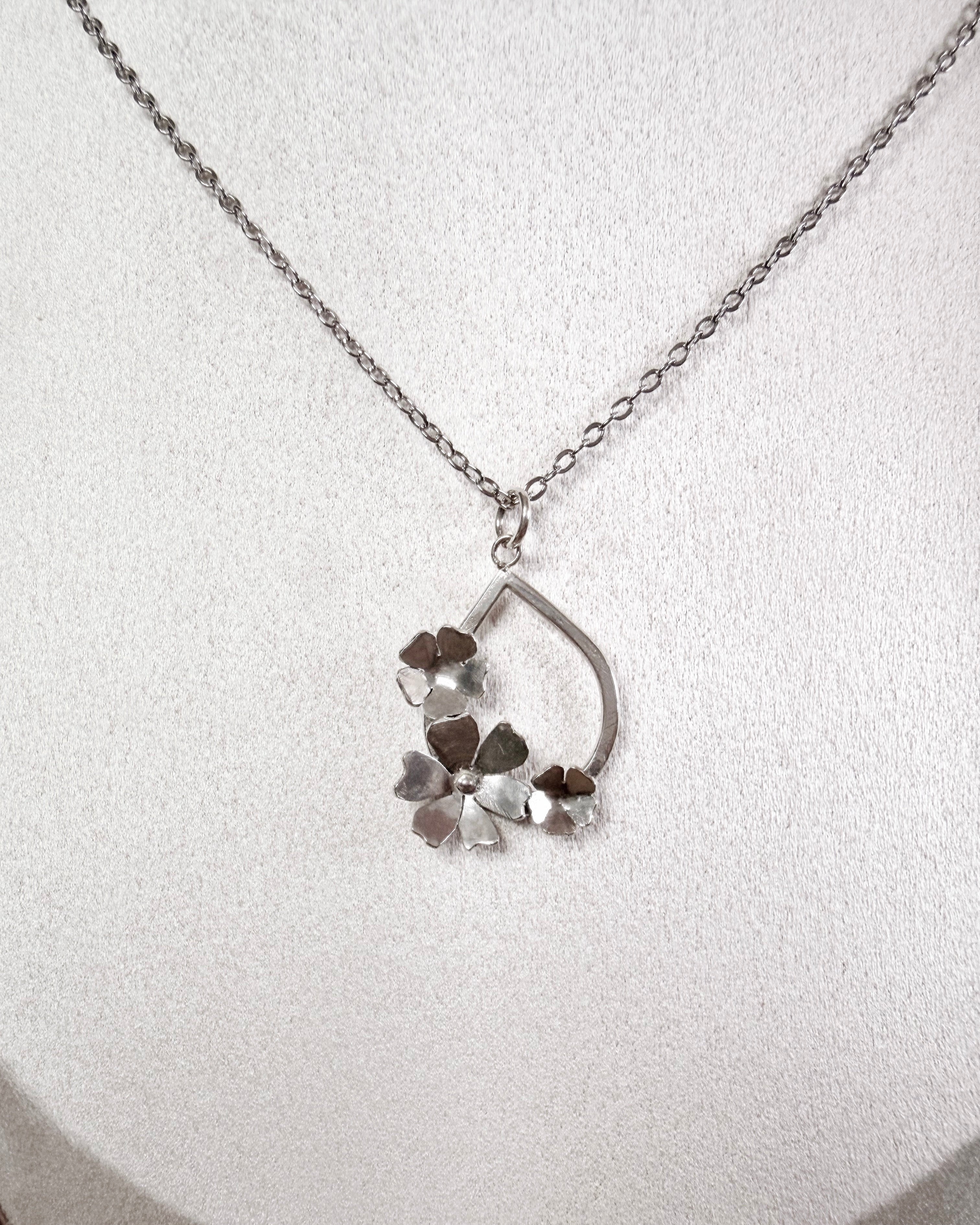 A silver necklace with a delicate chain and a pendant featuring a teardrop-shaped frame and three small flower-like charms, displayed on a textured white surface.