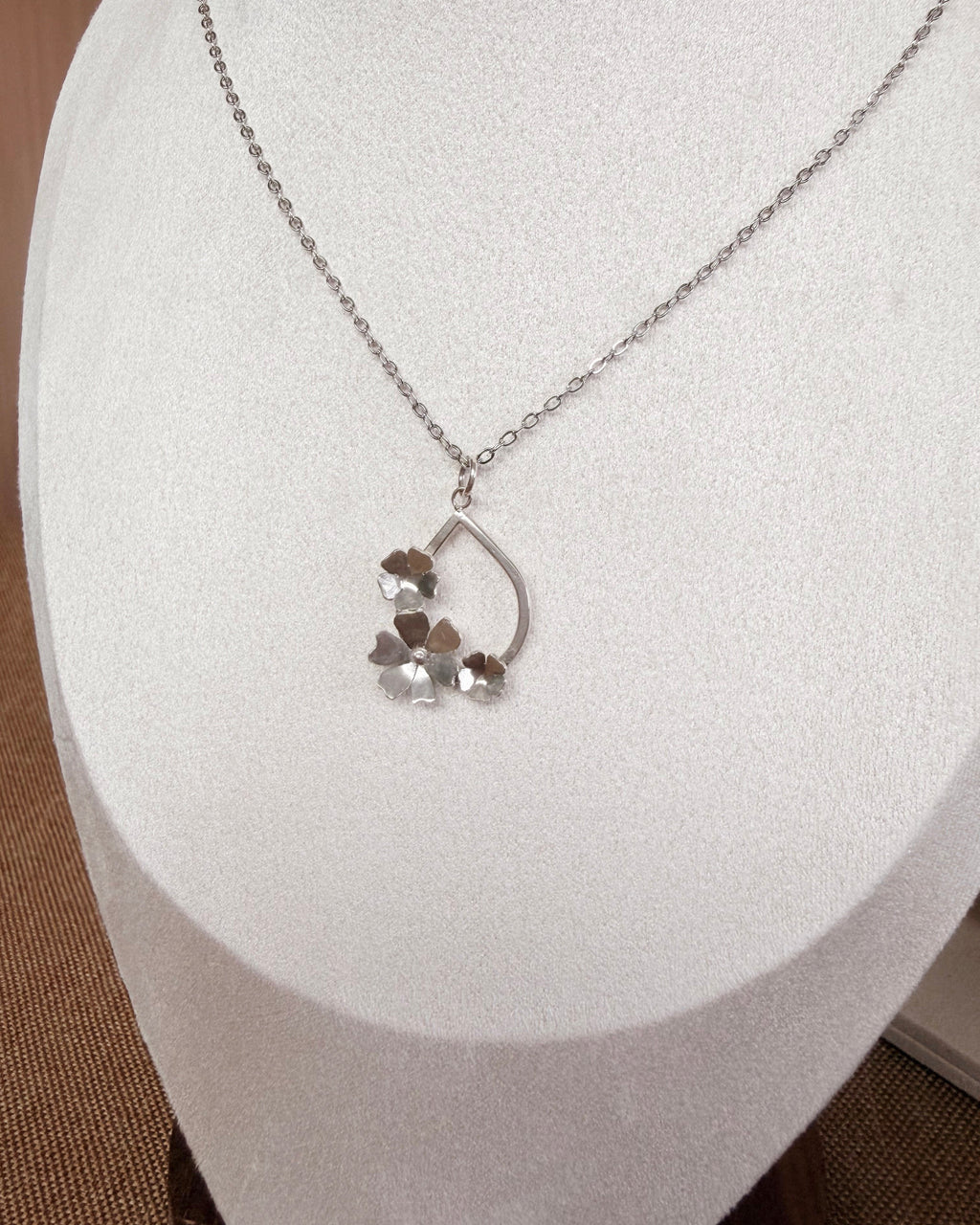 A silver necklace with a delicate chain and a small pendant featuring three small flowers, displayed on a white jewelry bust.