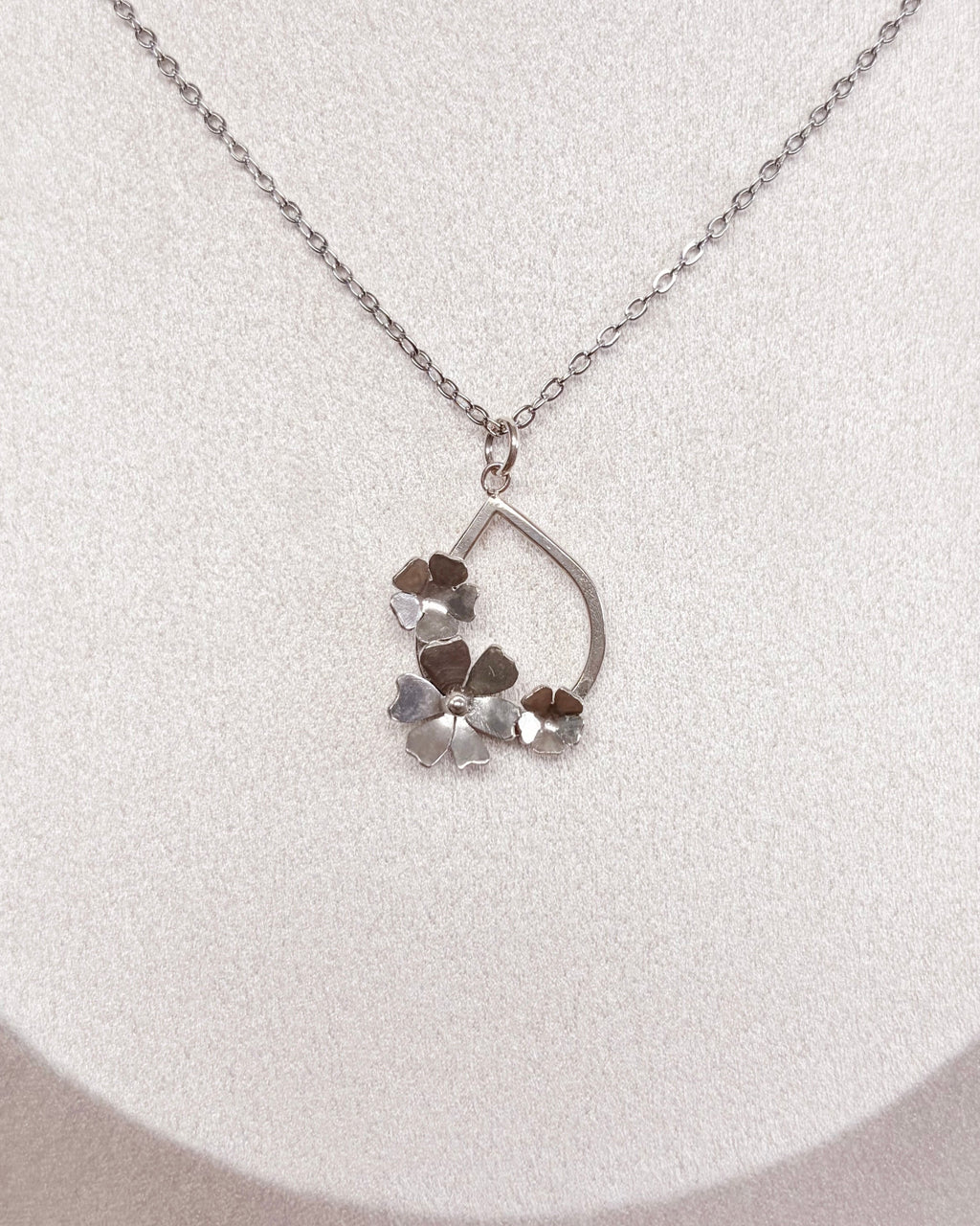 A delicate silver chain necklace with a small floral pendant, displayed on a white fabric jewelry bust.