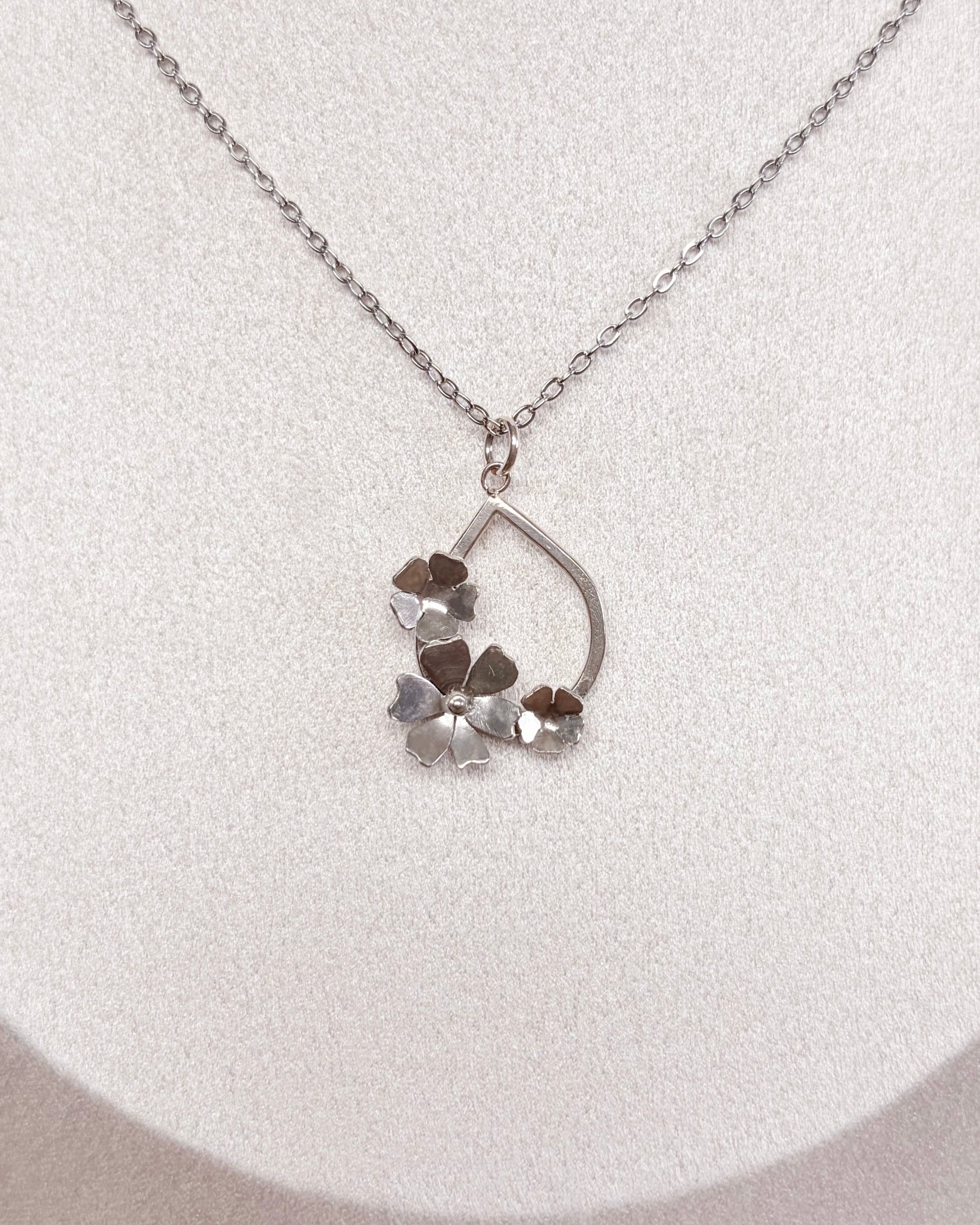 A delicate silver chain necklace with a small floral pendant, displayed on a white fabric jewelry bust.