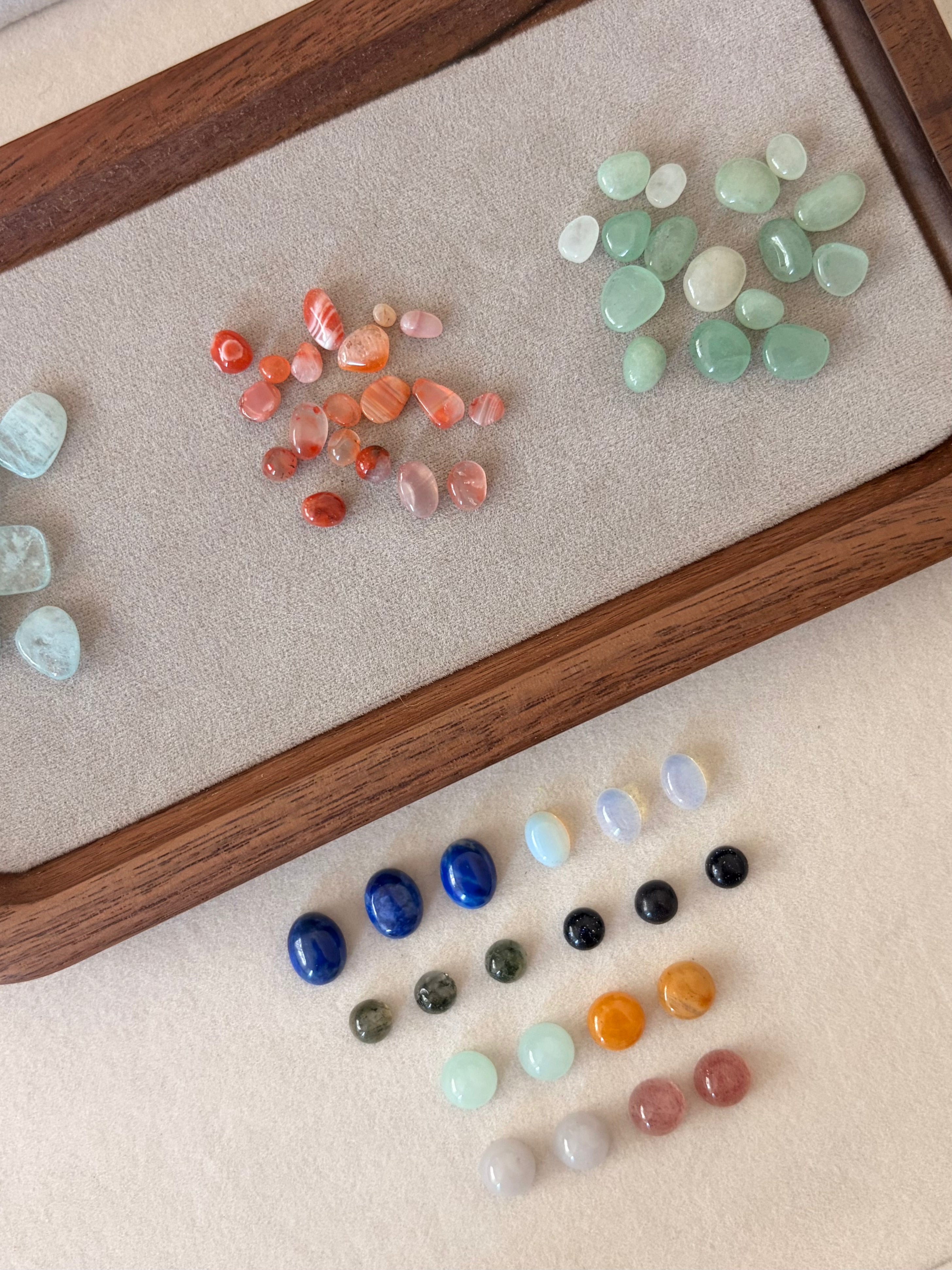 A collection of polished tumbled gemstones and crystals arranged on a beige fabric surface next to a wooden tray. The stones include various colors such as light green, orange, blue, black, white, and pink, some larger smooth teardrop shapes and others small round beads.