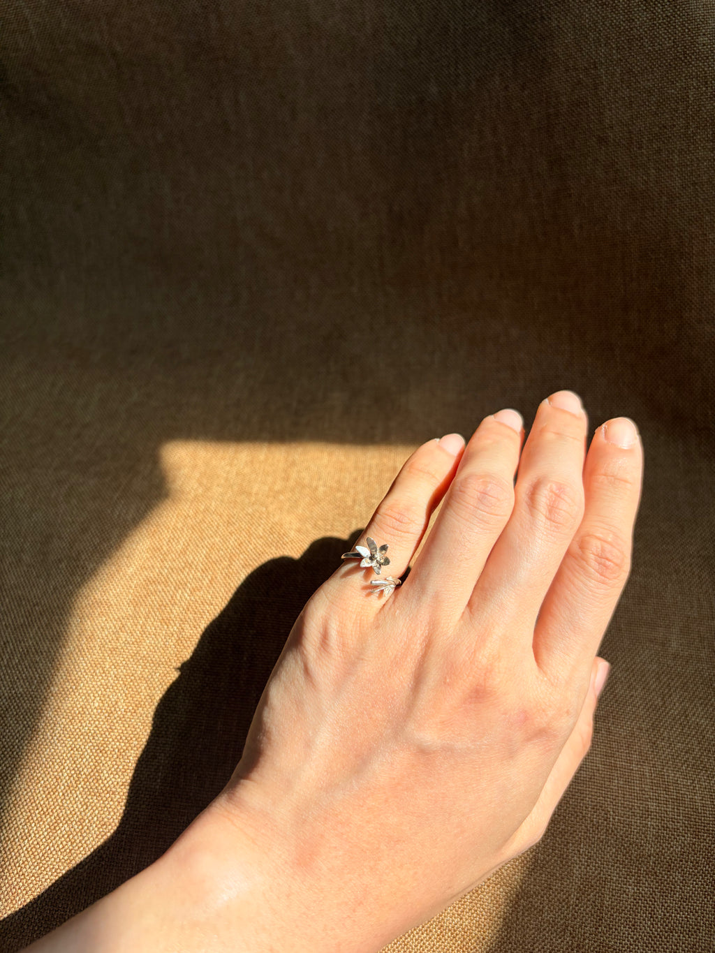 A silver ring with a jasmine floral design worn on a person's hand, set against a dark brown textured background with dramatic sunlight and shadows.
