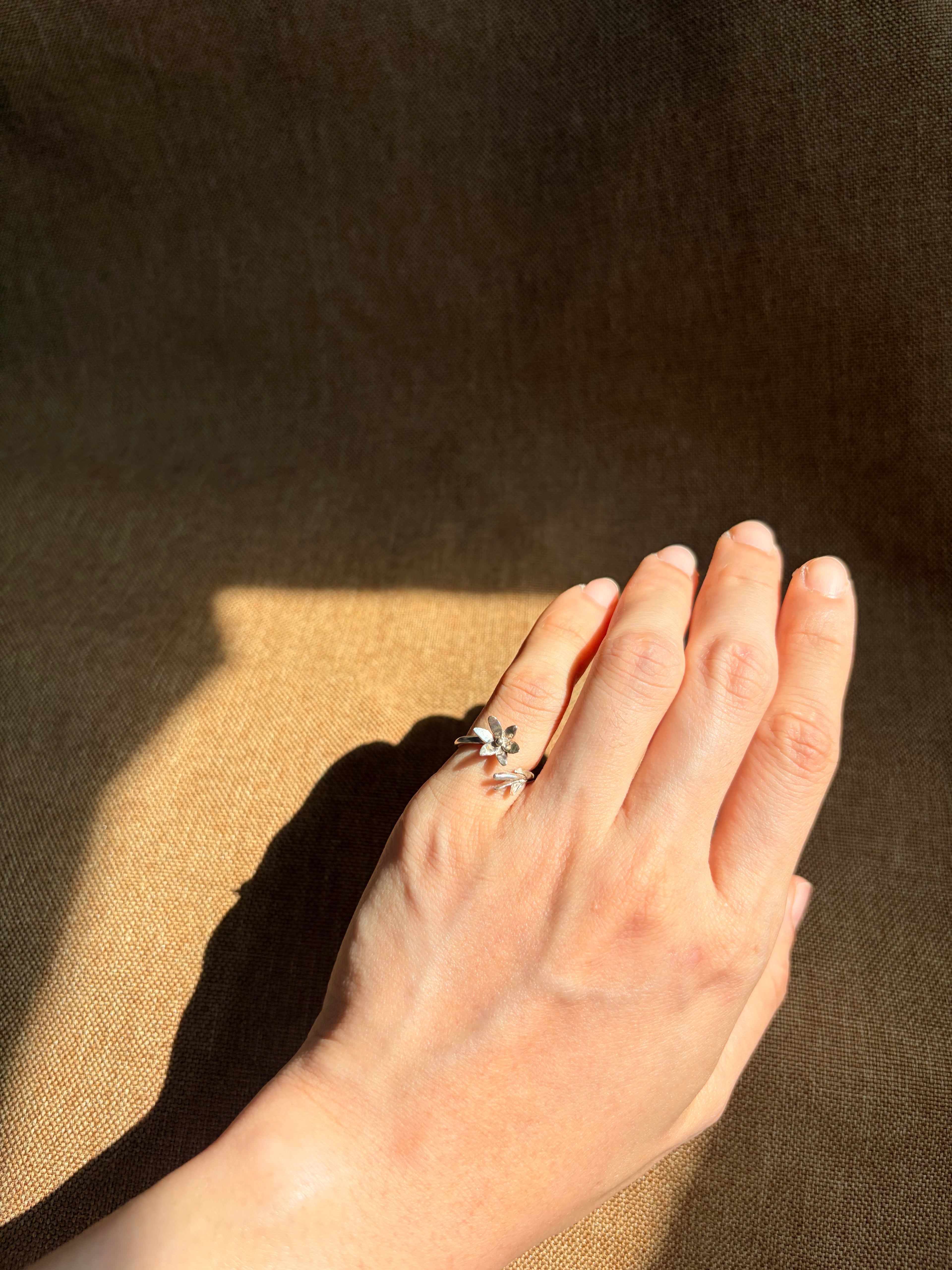 A silver ring with a jasmine floral design worn on a person's hand, set against a dark brown textured background with dramatic sunlight and shadows.