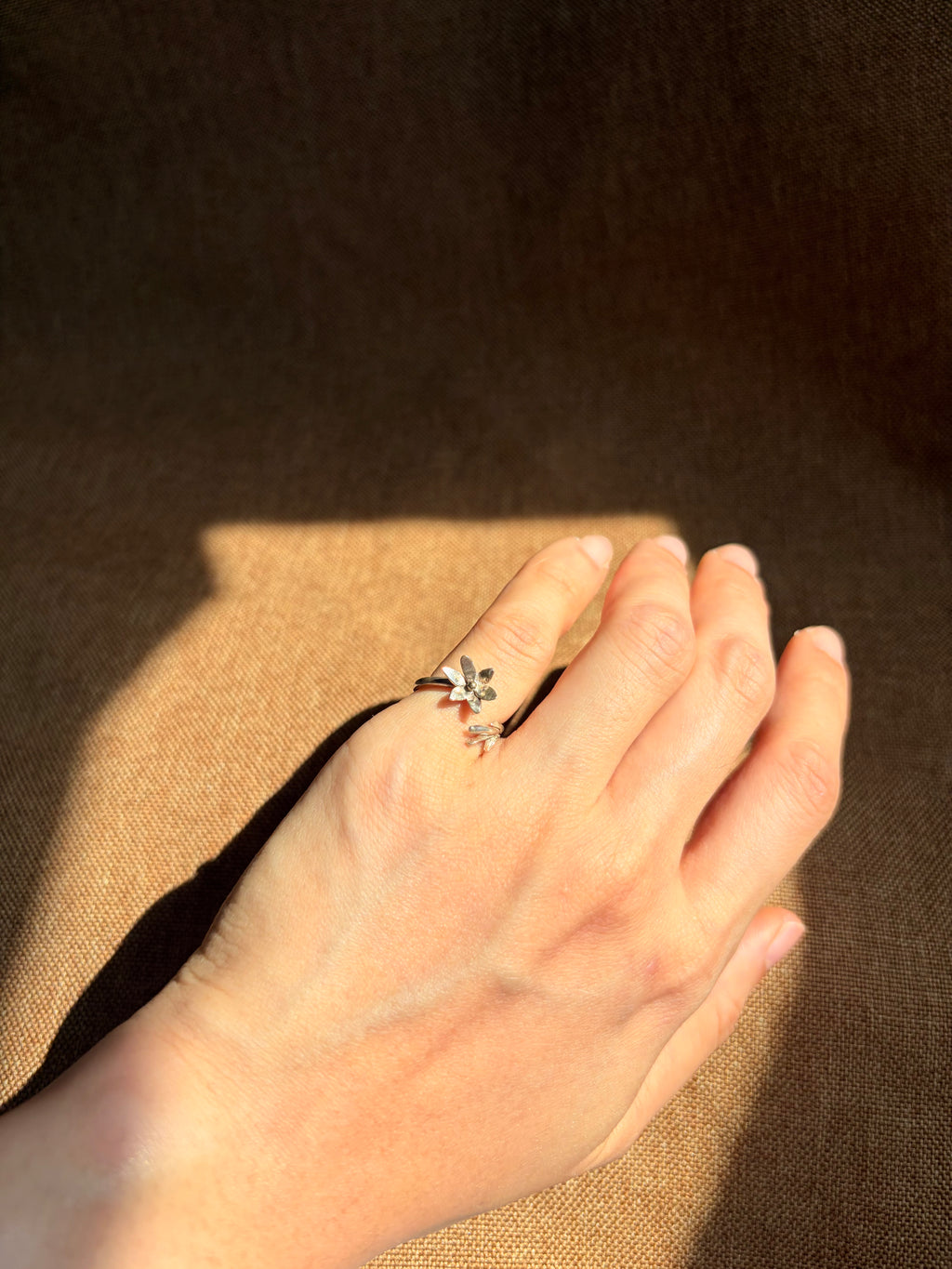 A delicate silver ring with small floral details, worn on the pinky finger of a hand resting against a textured brown background.