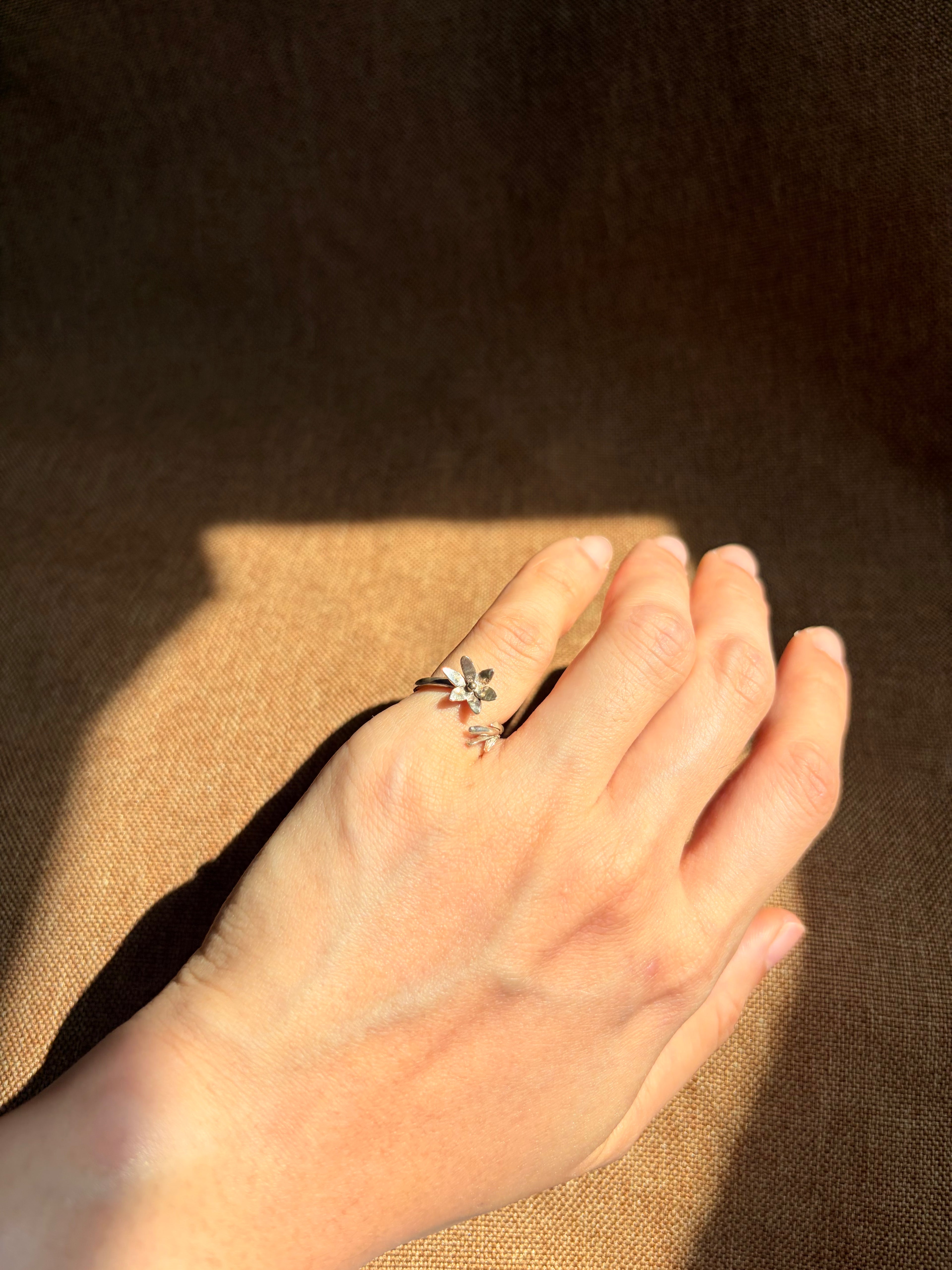 A delicate silver ring with small floral details, worn on the pinky finger of a hand resting against a textured brown background.