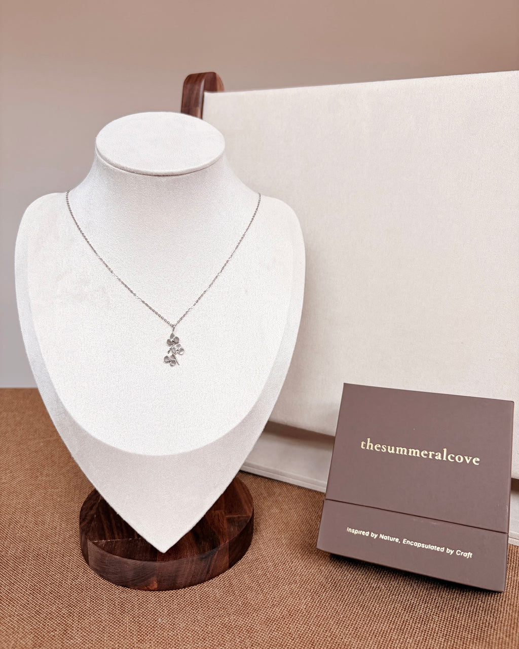 A silver-toned orchid pendant necklace displayed on a white jewelry bust, with a small brown brand box labeled 'thesummeralcove' beside it on a textured beige surface.