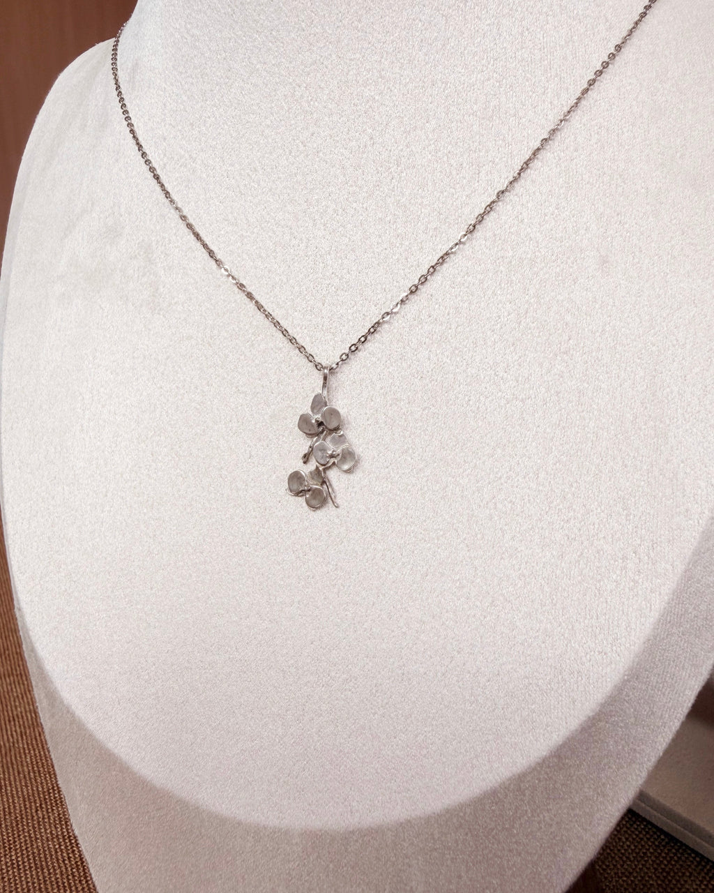 A delicate silver chain necklace with a small floral pendant displayed on a white jewelry bust.