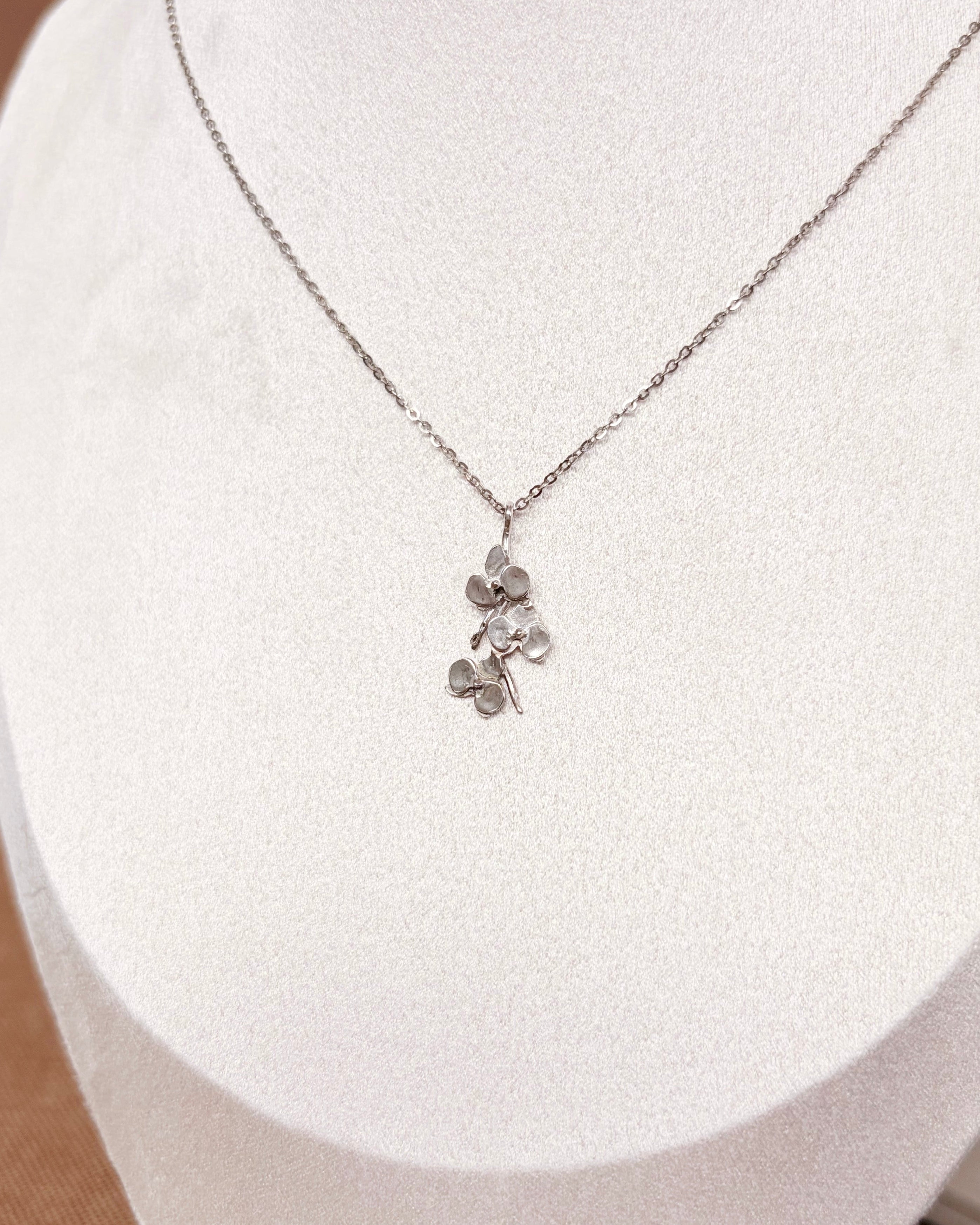 A delicate silver chain necklace with a small silver orchid pendant, displayed on a white fabric jewelry bust.